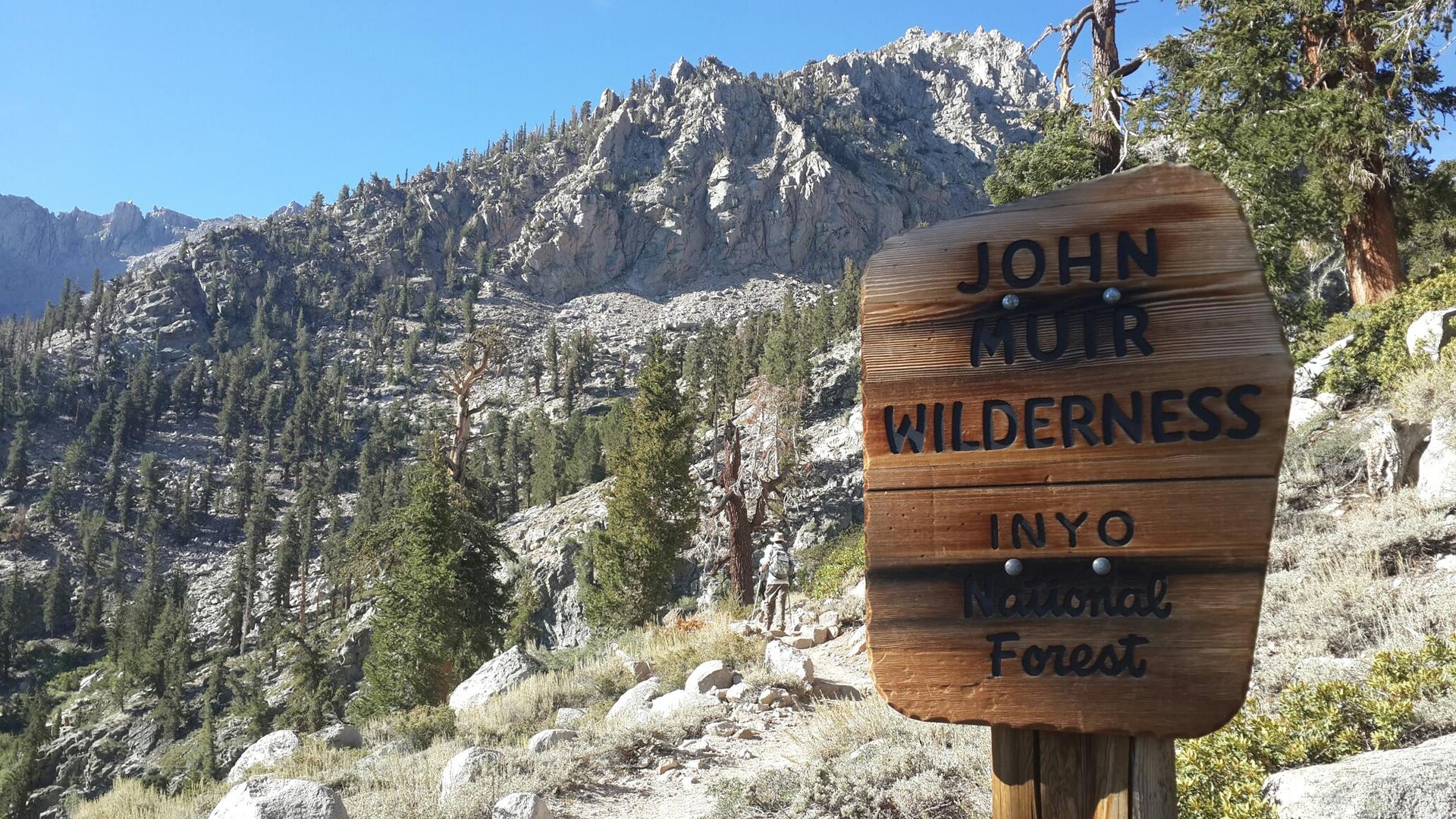 Wooden signpost on a rocky mountain trail with a hiker and peaks in background.