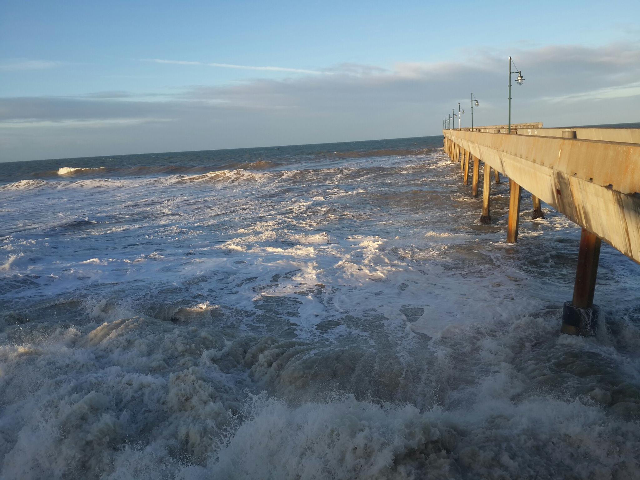 Rough ocean waves crash against a concrete pier under a golden sky.