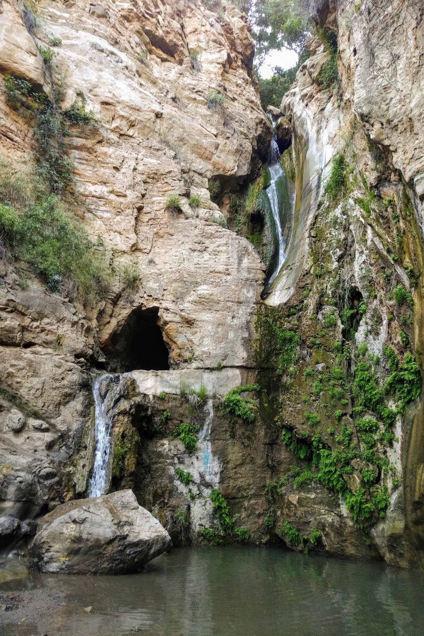 Two waterfalls cascade into a dark pool within a rocky, green canyon.