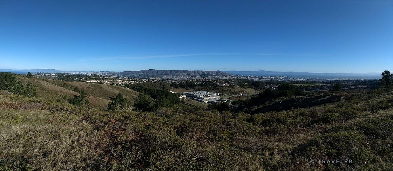 Panoramic view of rolling hills, a distant town, and mountains under clear sky.