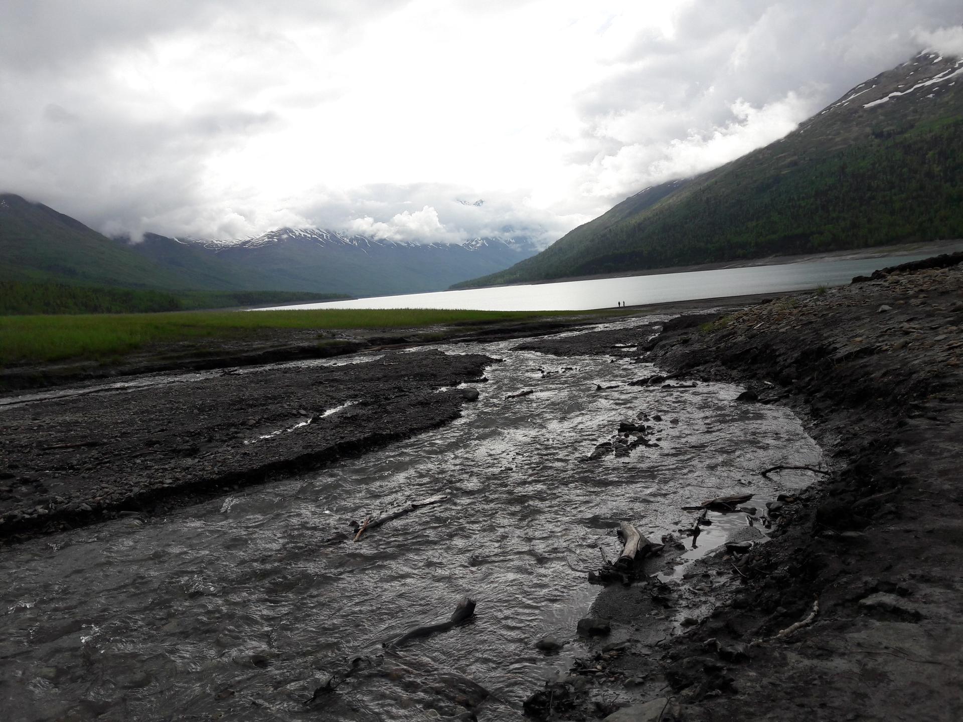 Muddy stream flows into a lake, two distant figures under cloudy skies.
