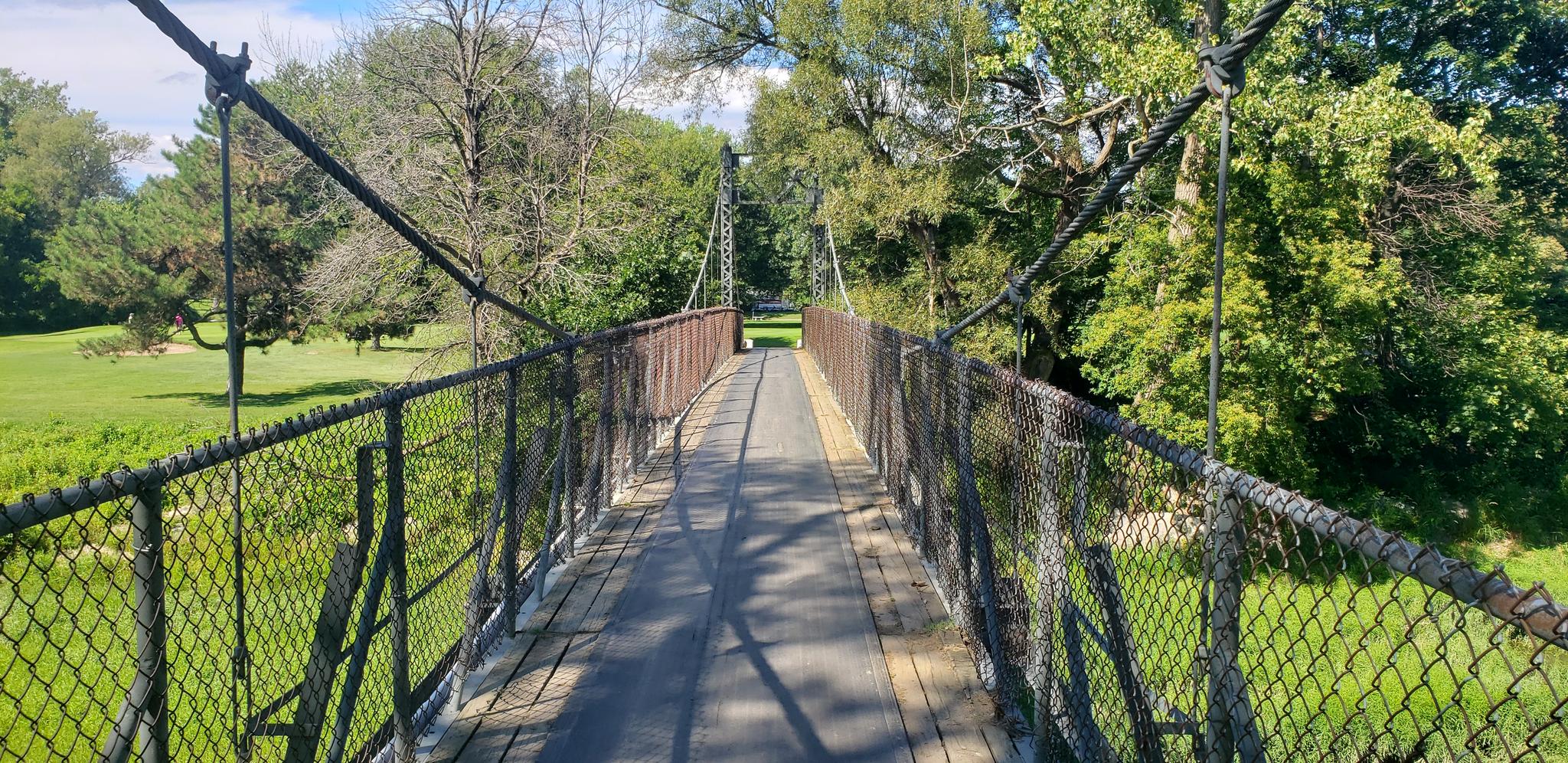 Wooden suspension bridge with chain link fences over green grass and trees.