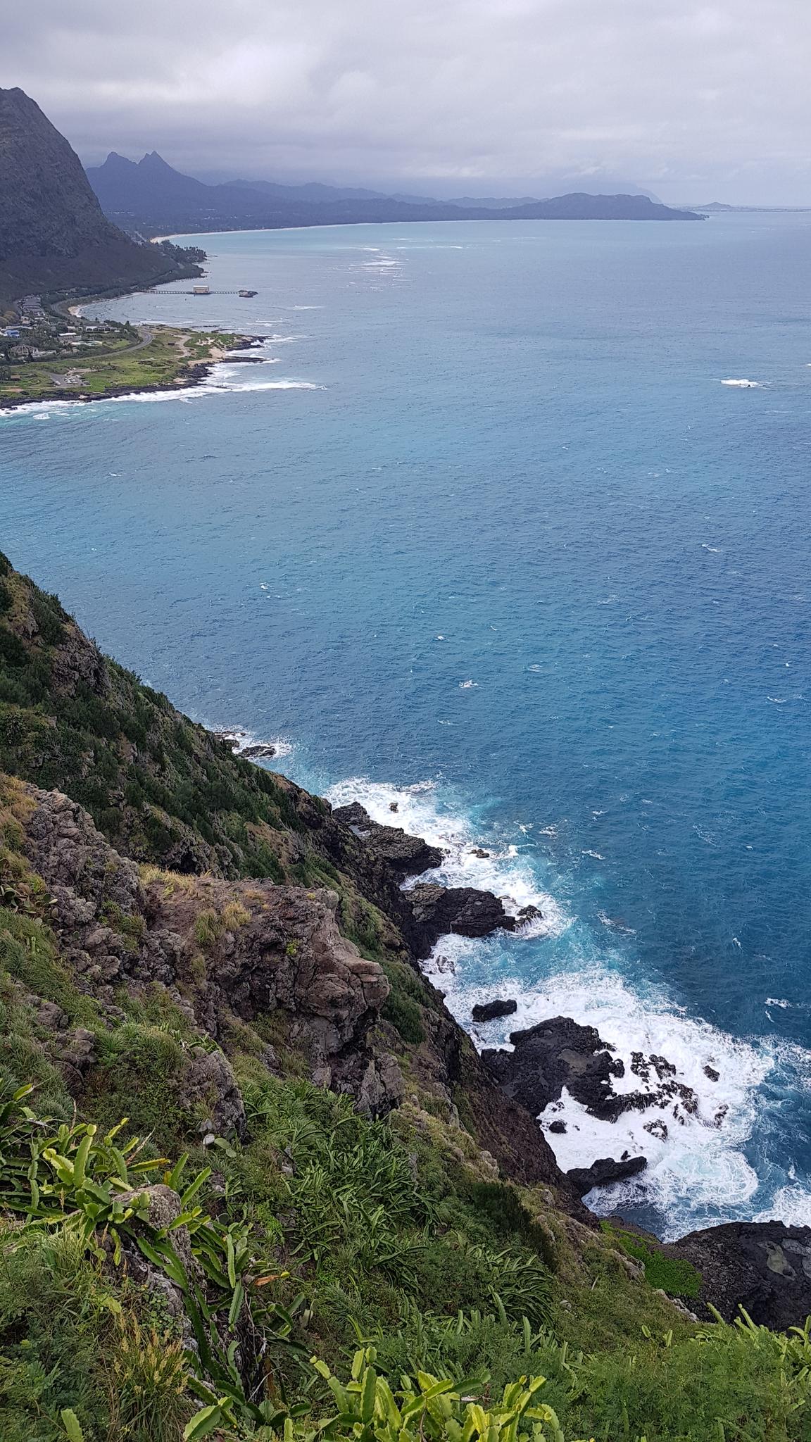 Steep green cliffside overlooking a blue ocean with white waves and distant coastline.
