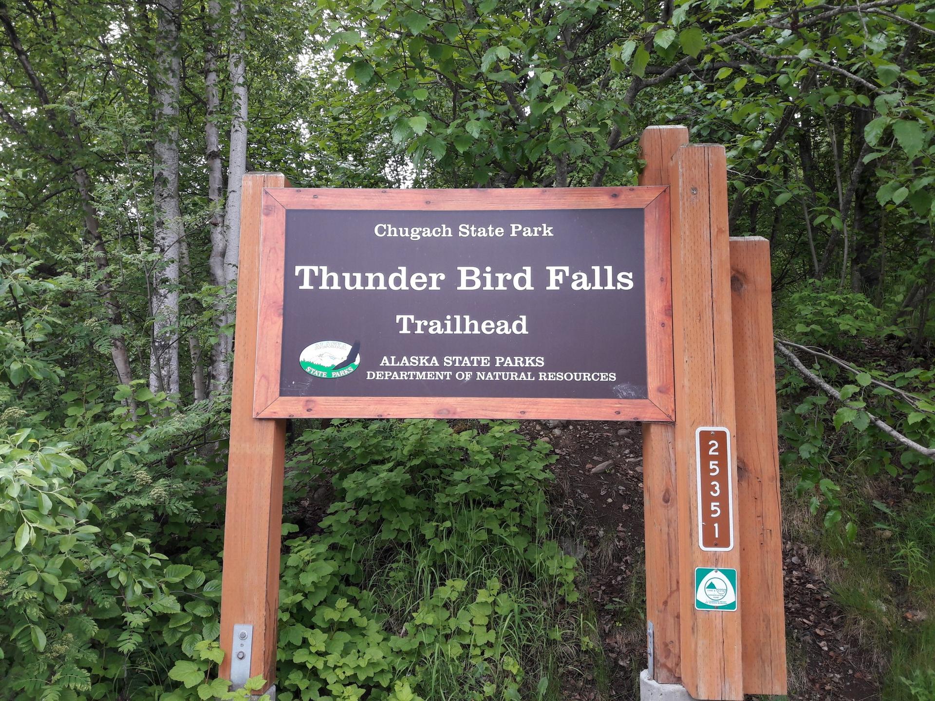 A wooden trailhead sign stands amidst green trees and undergrowth.