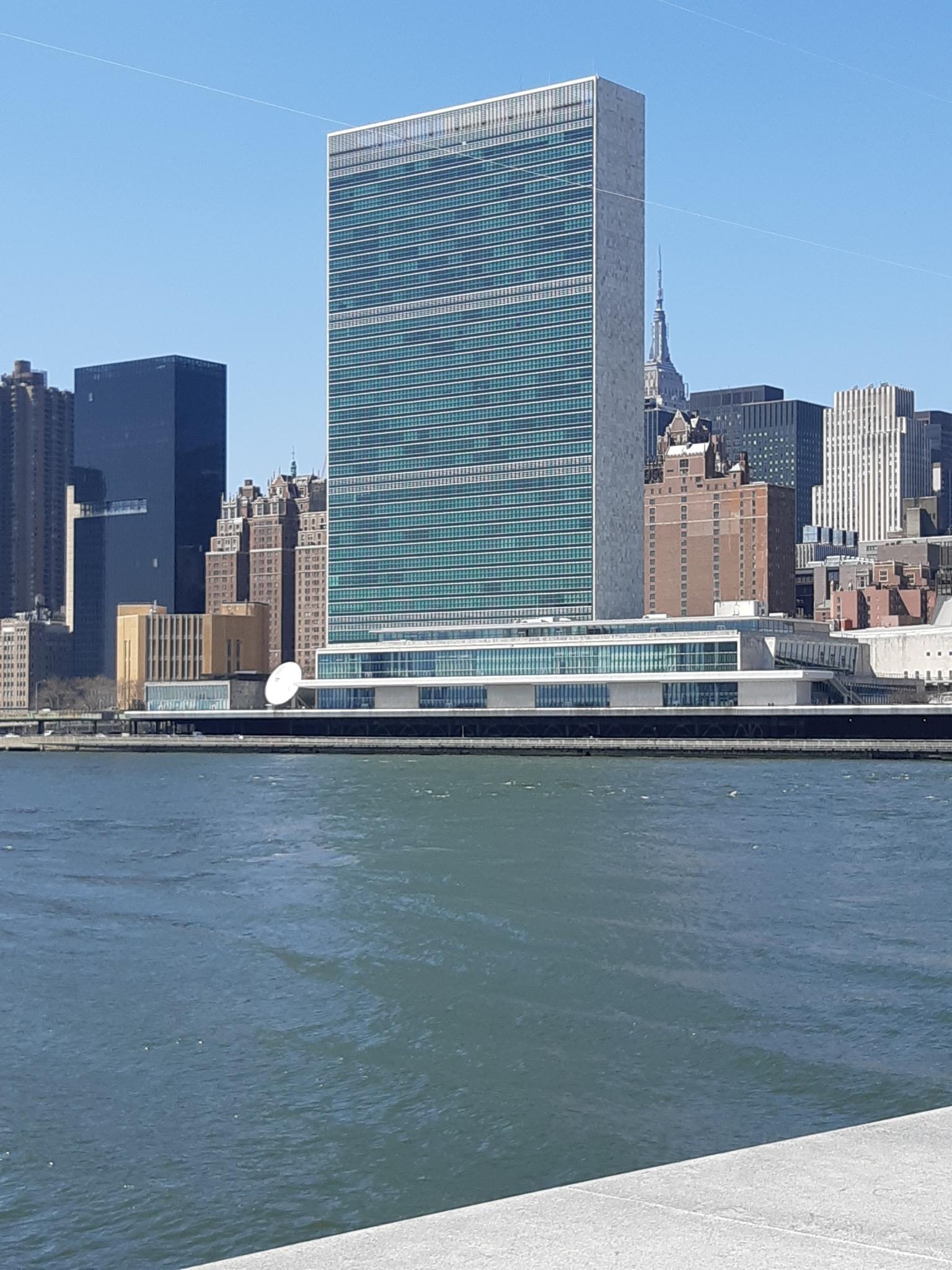 A prominent tall, rectangular building stands across a river on a sunny day.
