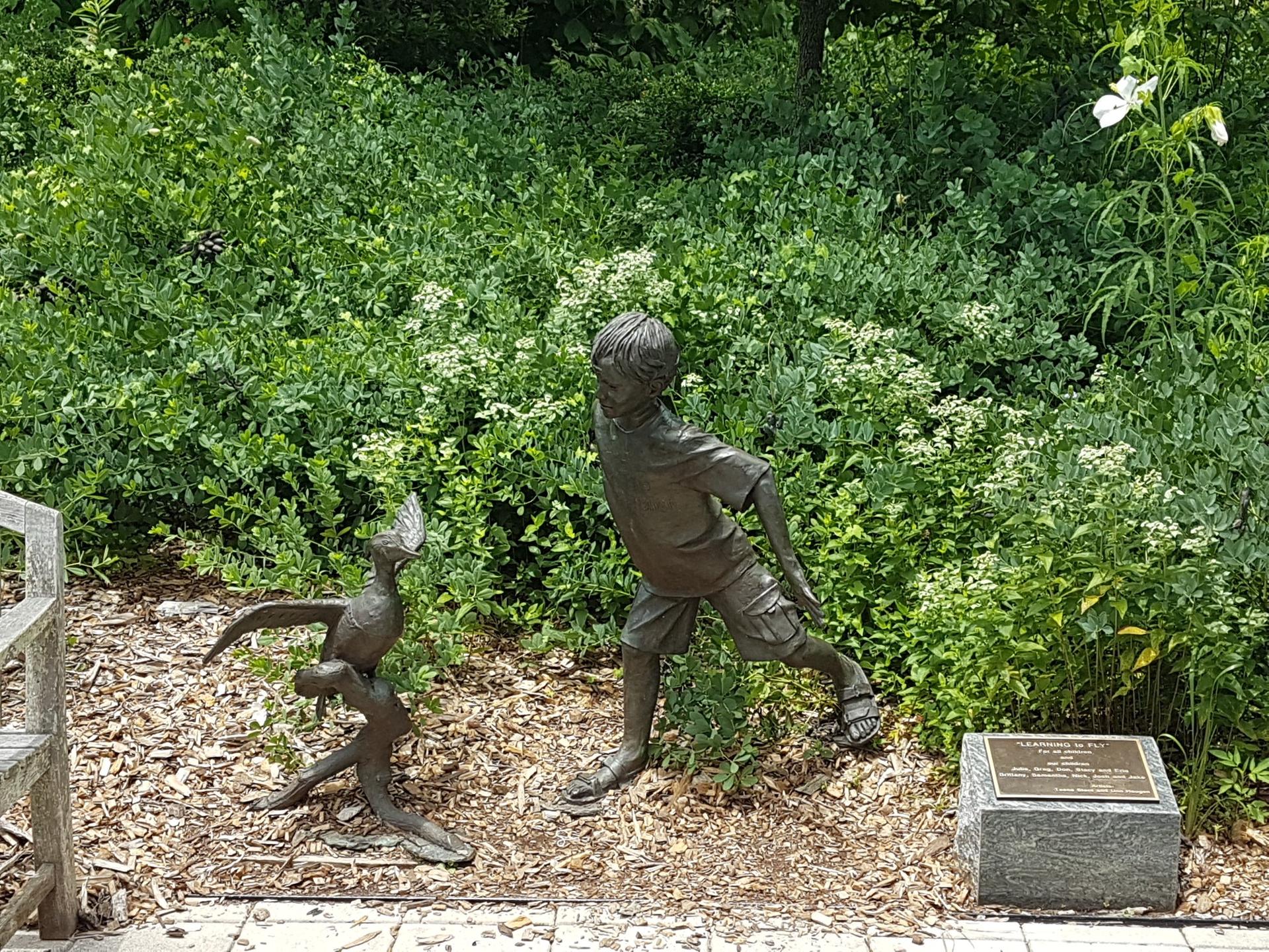 Bronze statues of a boy and a bird in a lush garden setting.