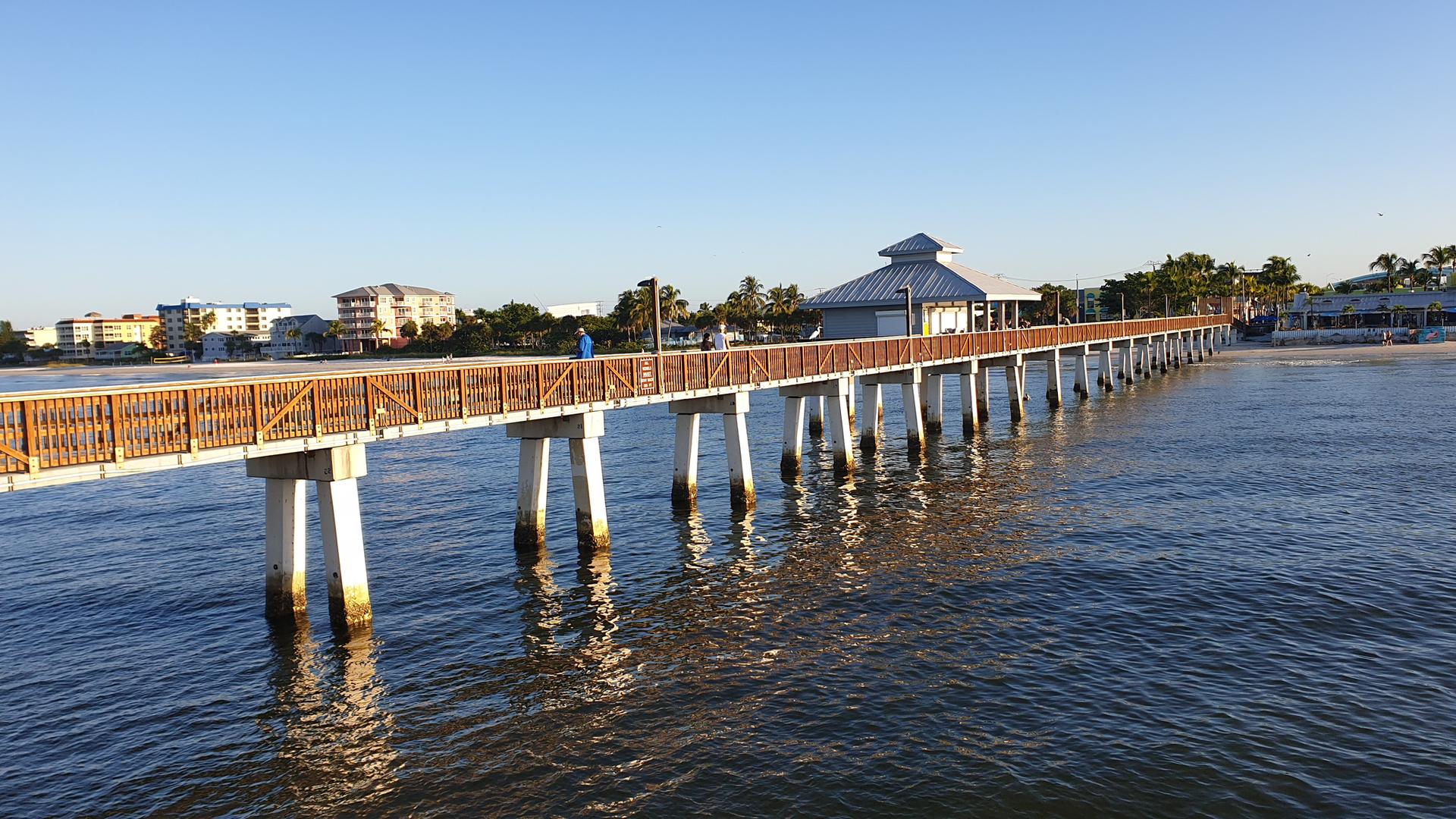 Long wooden pier extending over calm water with distant buildings and clear sky.