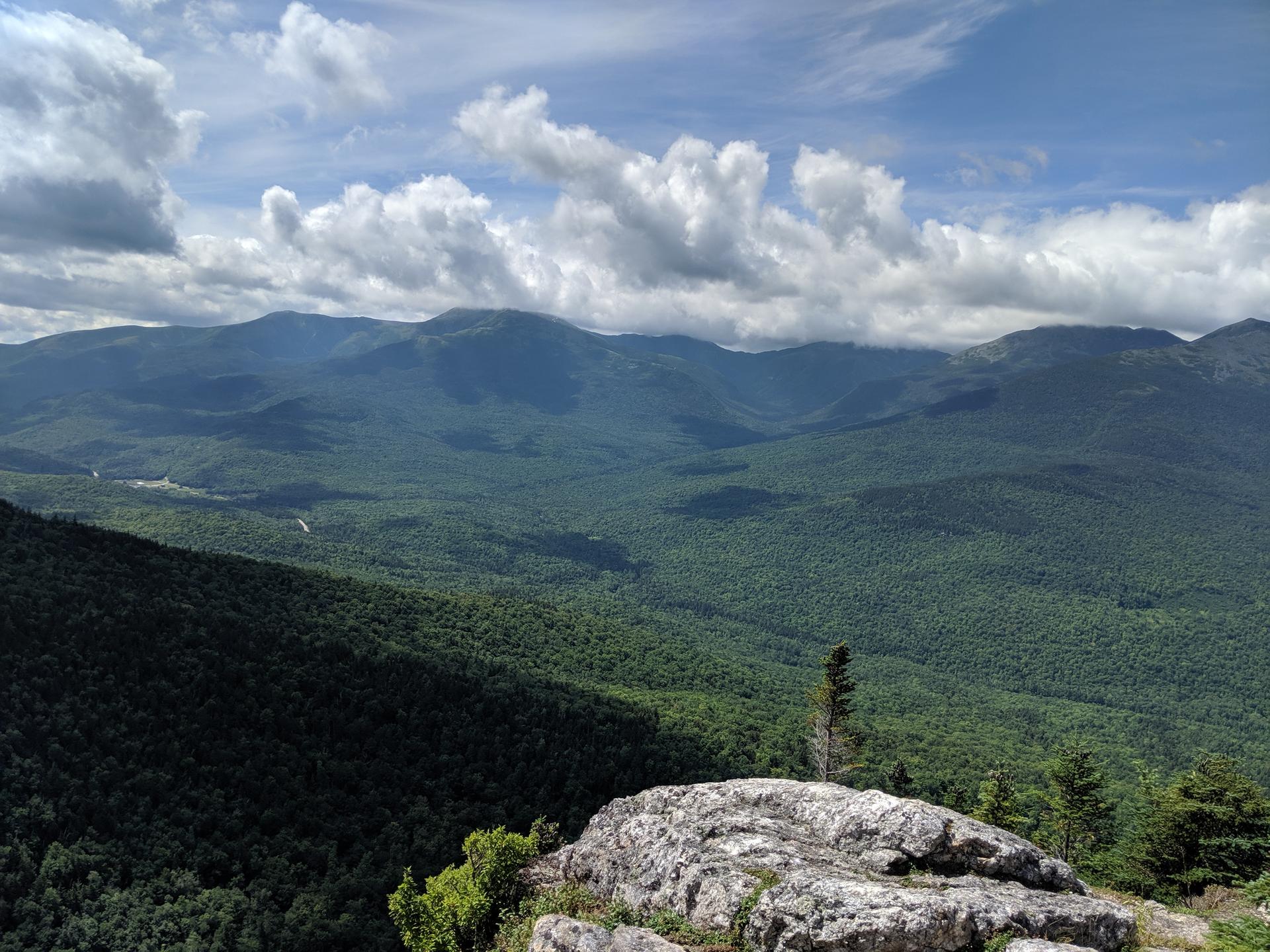 Vast green mountain landscape under a partly cloudy sky from a rocky viewpoint.