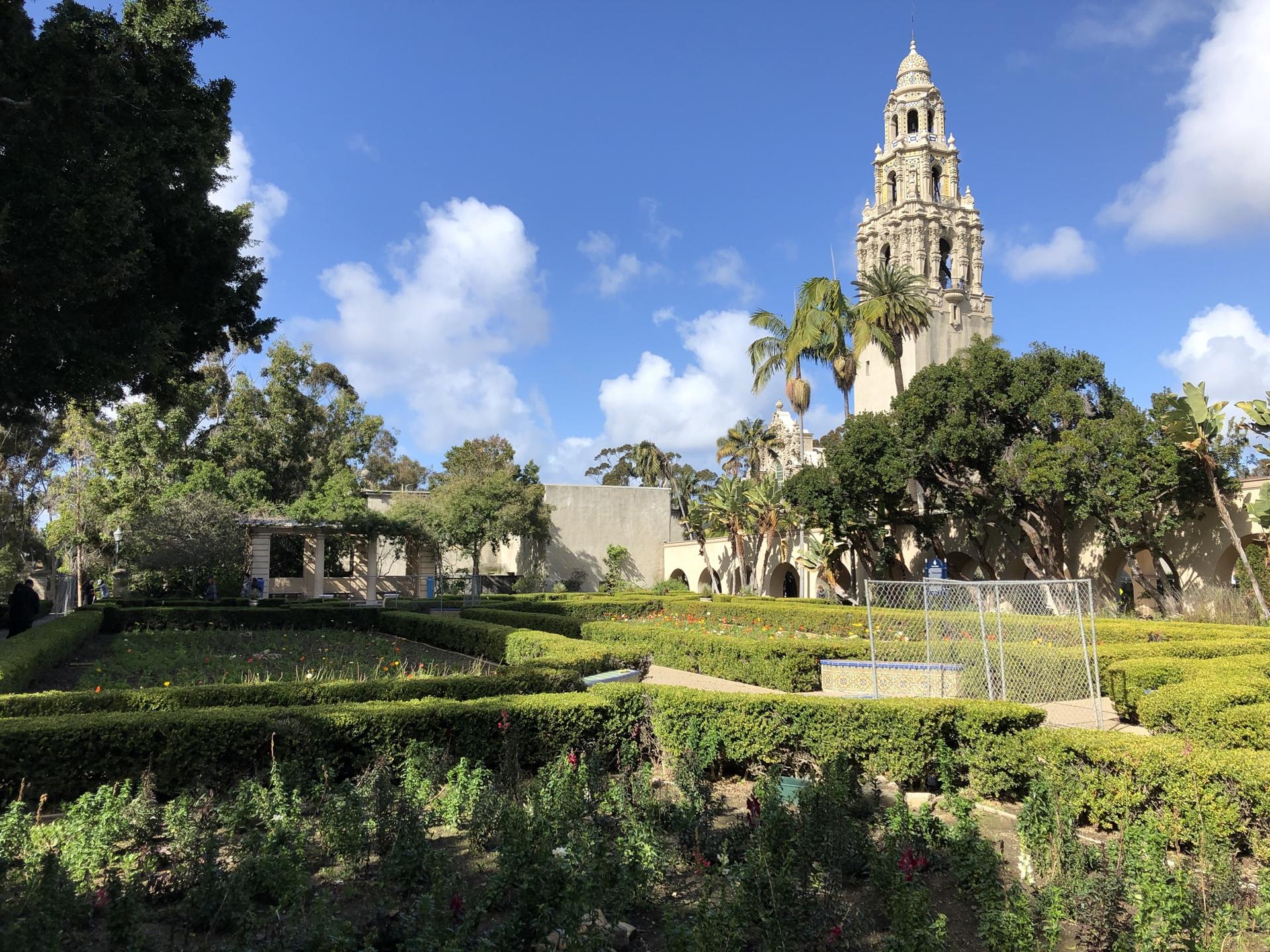 Ornate tower building behind a formal hedge garden under a bright sky.