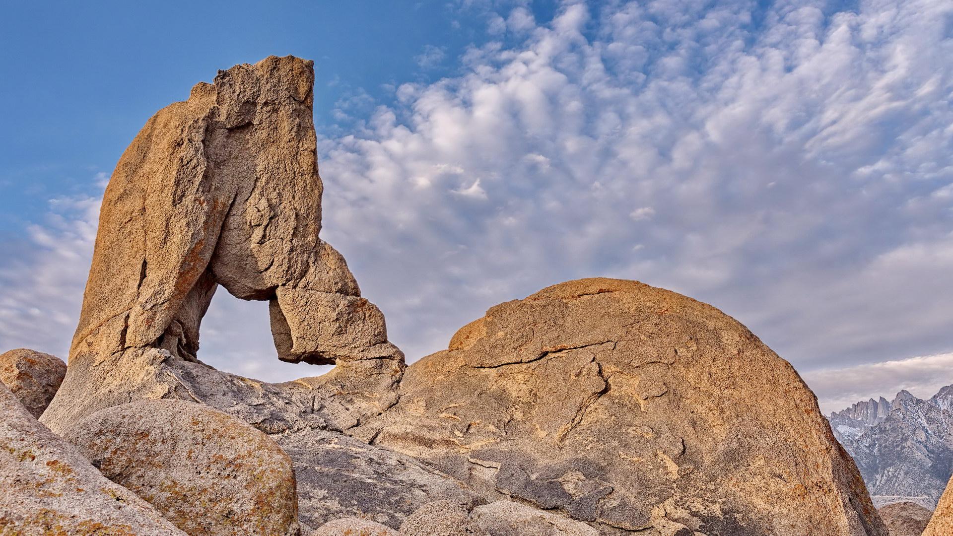 Dramatic rock arch formation against a cloudy blue sky and distant mountains.