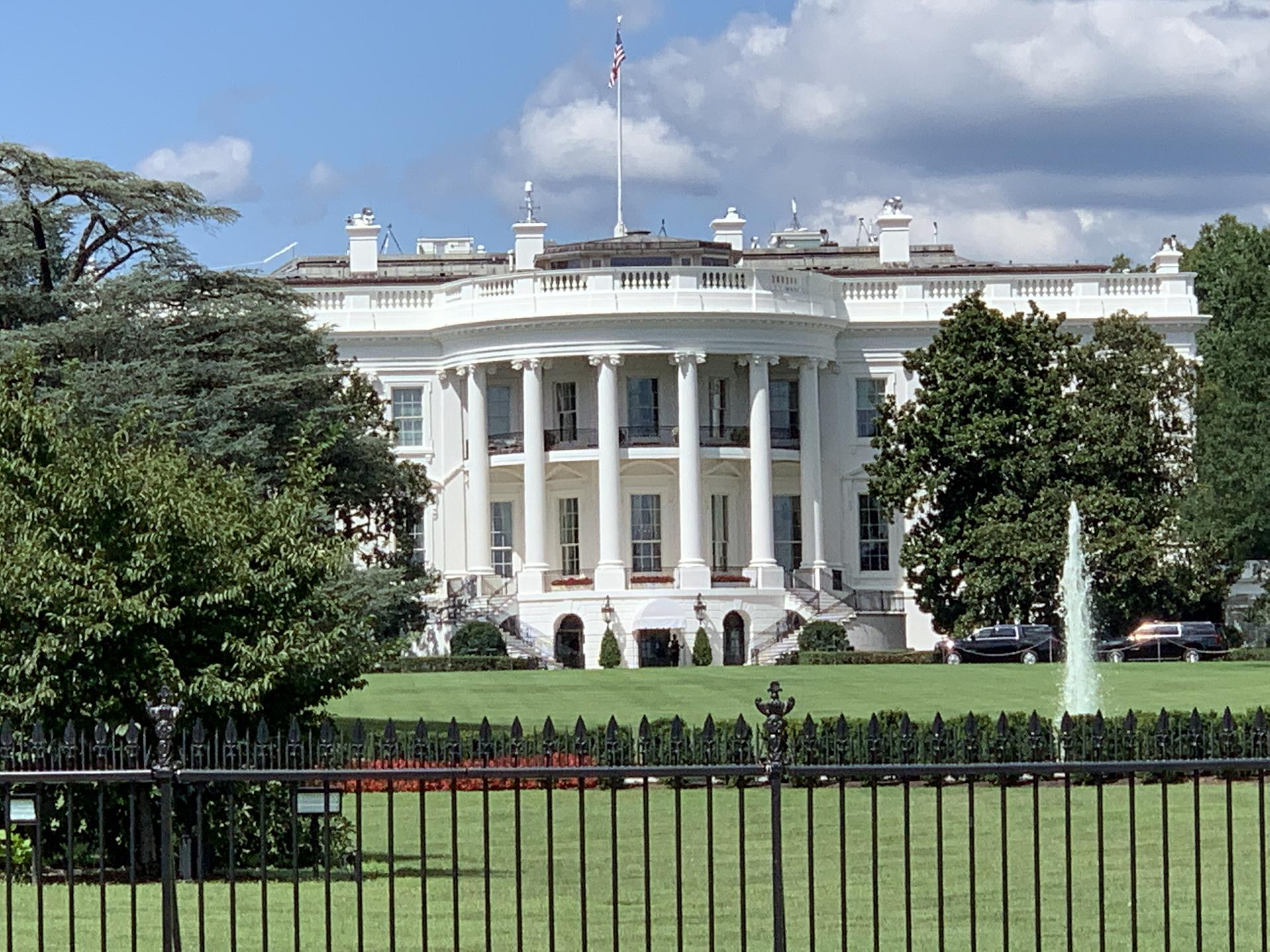 Grand white building with columns, green lawn, and fountain under blue sky.