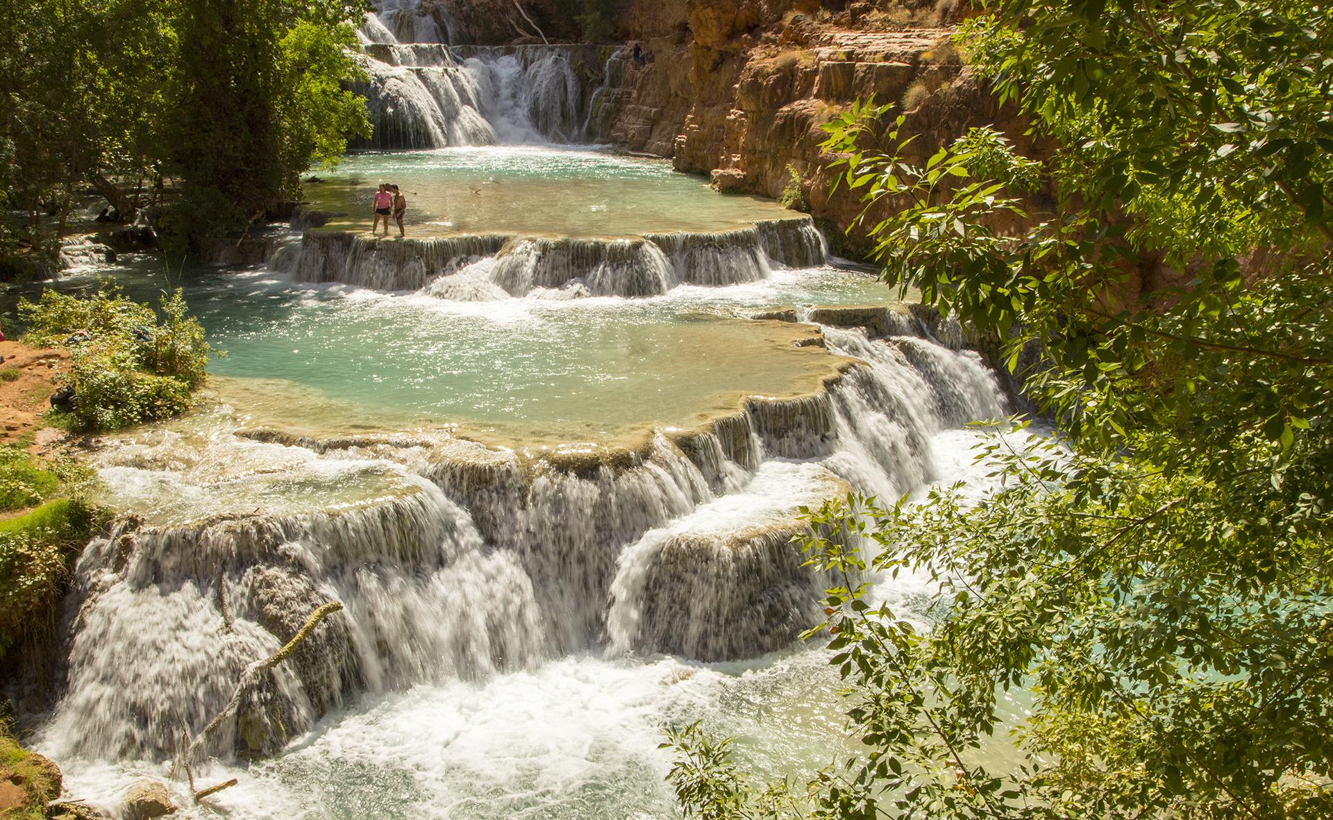 Two people stand in a tiered turquoise waterfall surrounded by green trees, sunny.