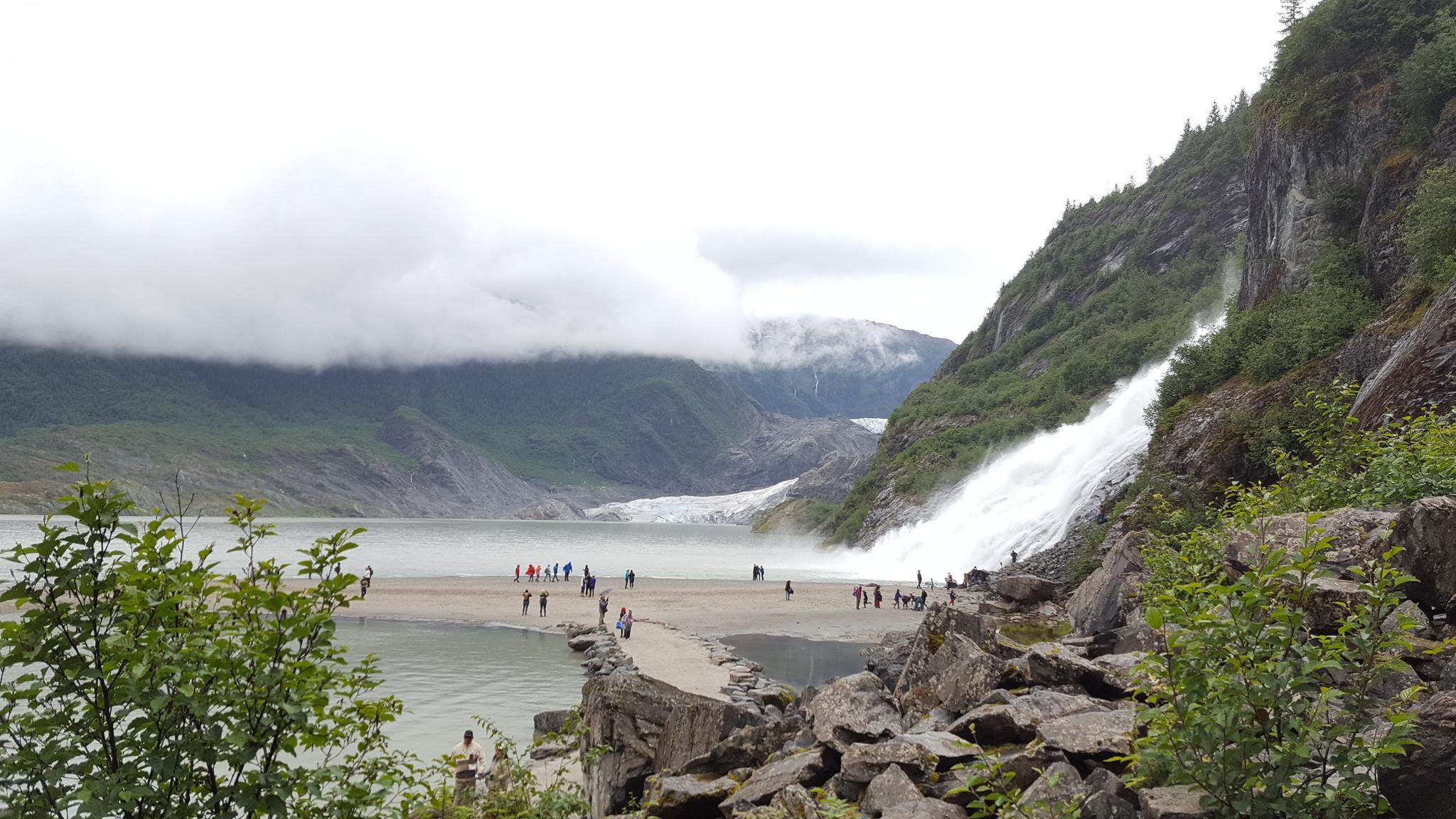 People gather near a powerful waterfall cascading into a lake with a distant glacier.