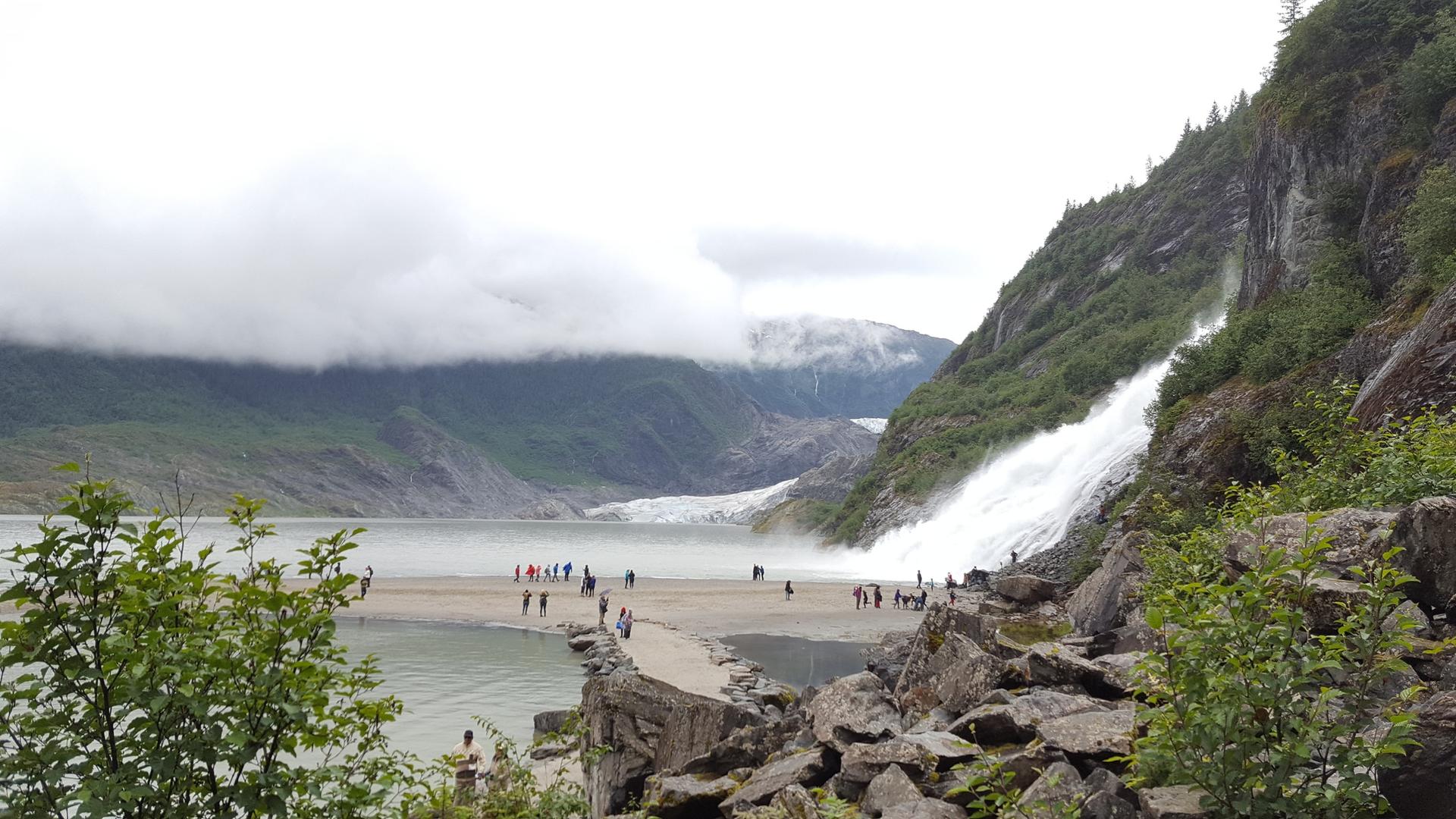 People gather near a powerful waterfall cascading into a lake with a distant glacier.