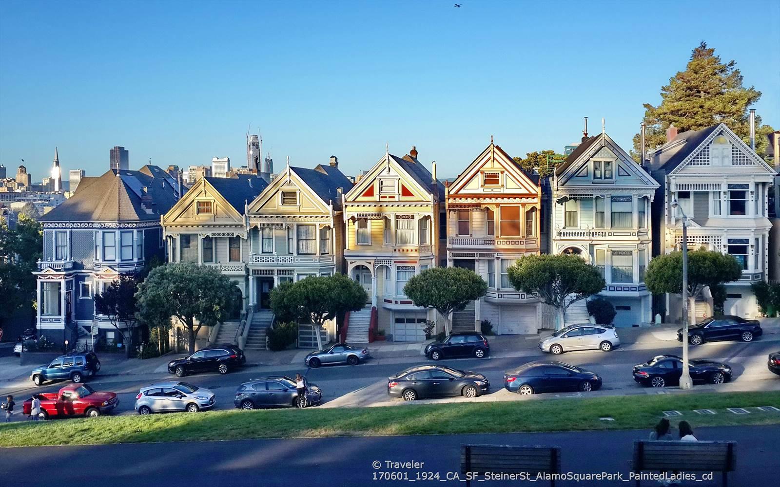 Row of colorful Victorian houses with cars parked on a street, under clear sky.