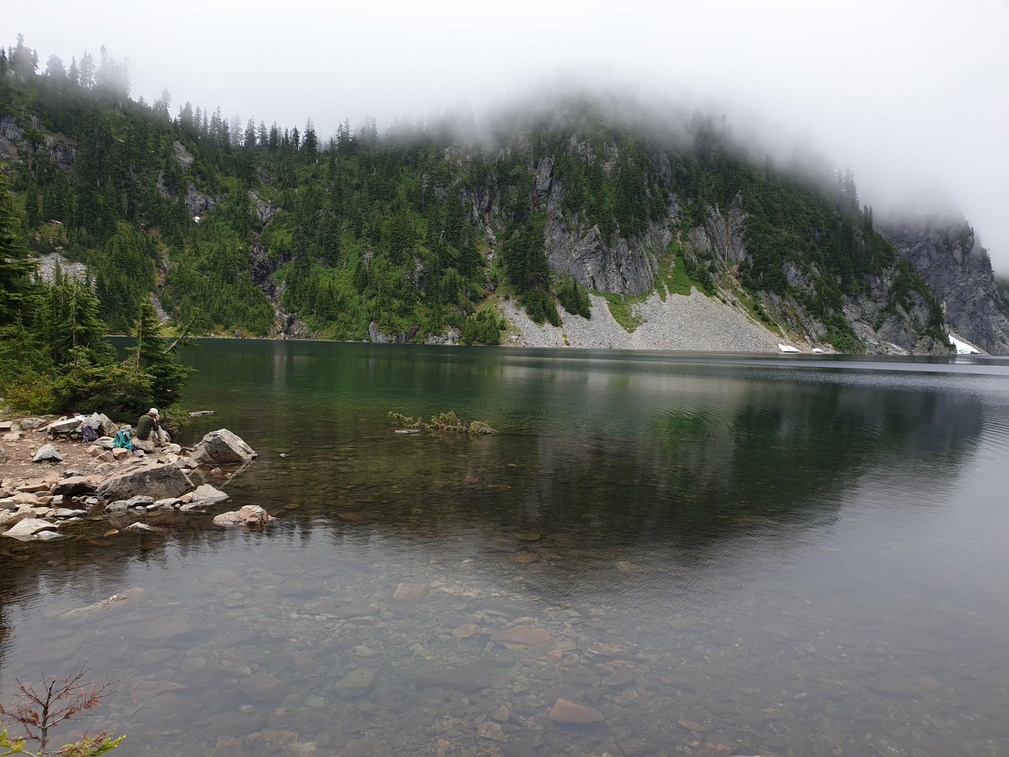 A person sits by a clear lake with misty, forested mountains under an overcast sky.