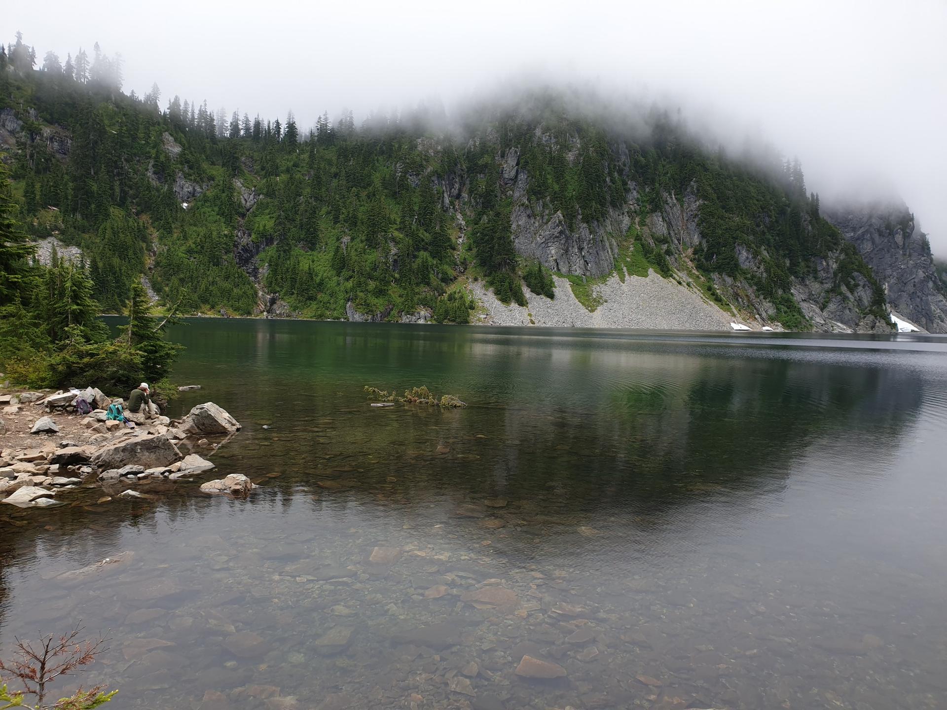 A person sits by a clear lake with misty, forested mountains under an overcast sky.