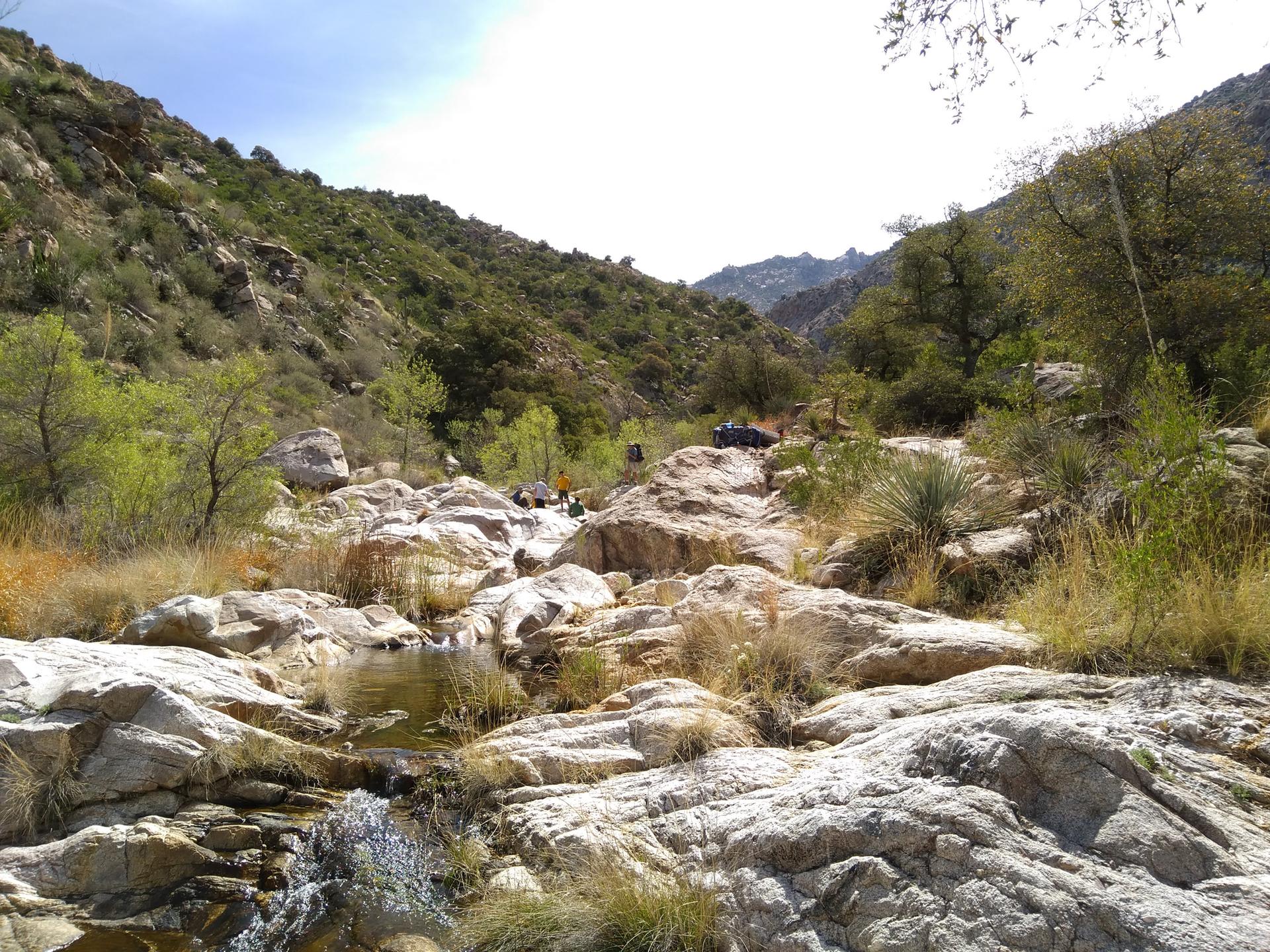 Rocky stream bed in a sunny canyon with green hillsides and distant people.