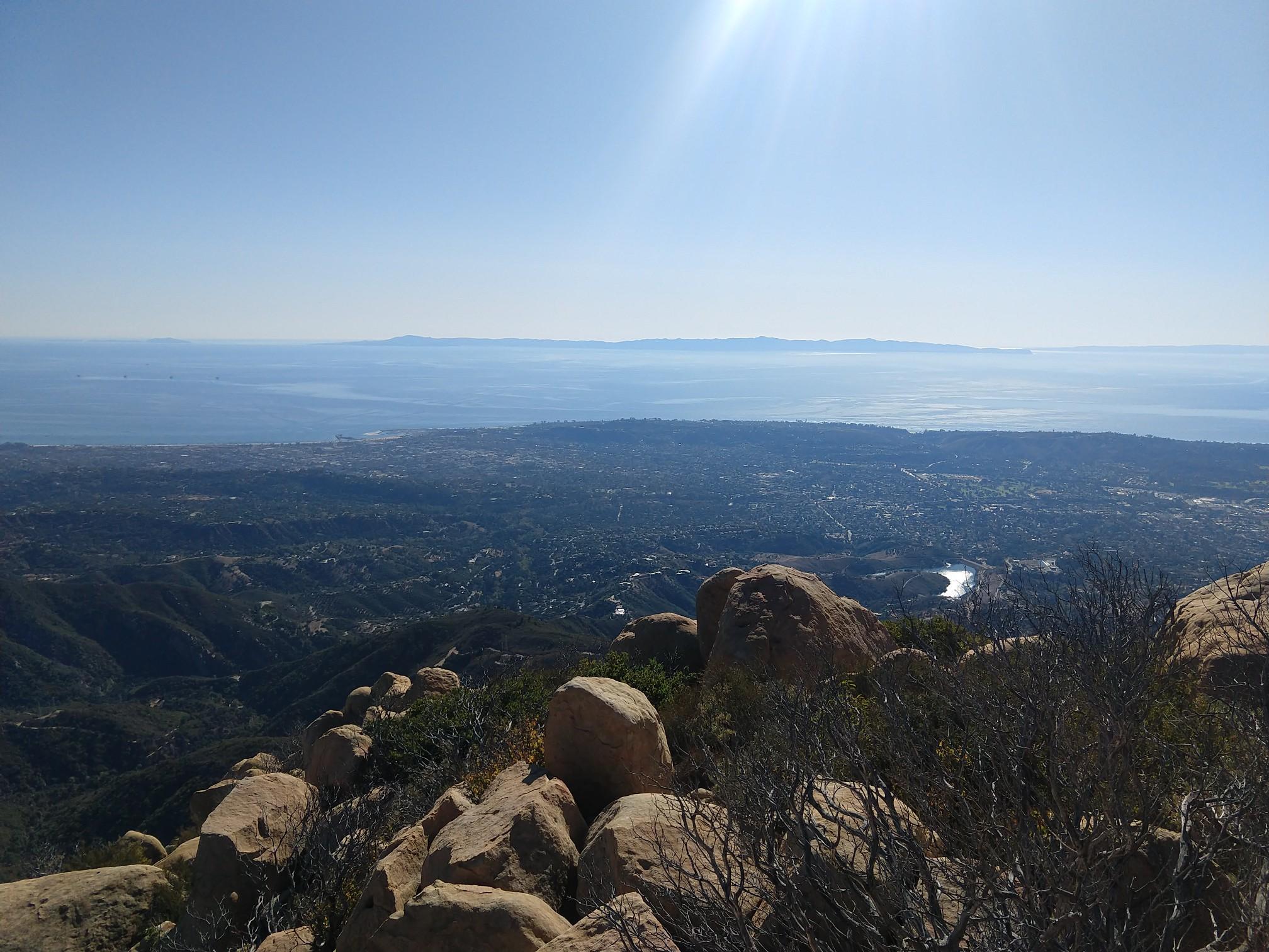 Sunny panoramic view from rocky peak overlooking a city, valley, and ocean.