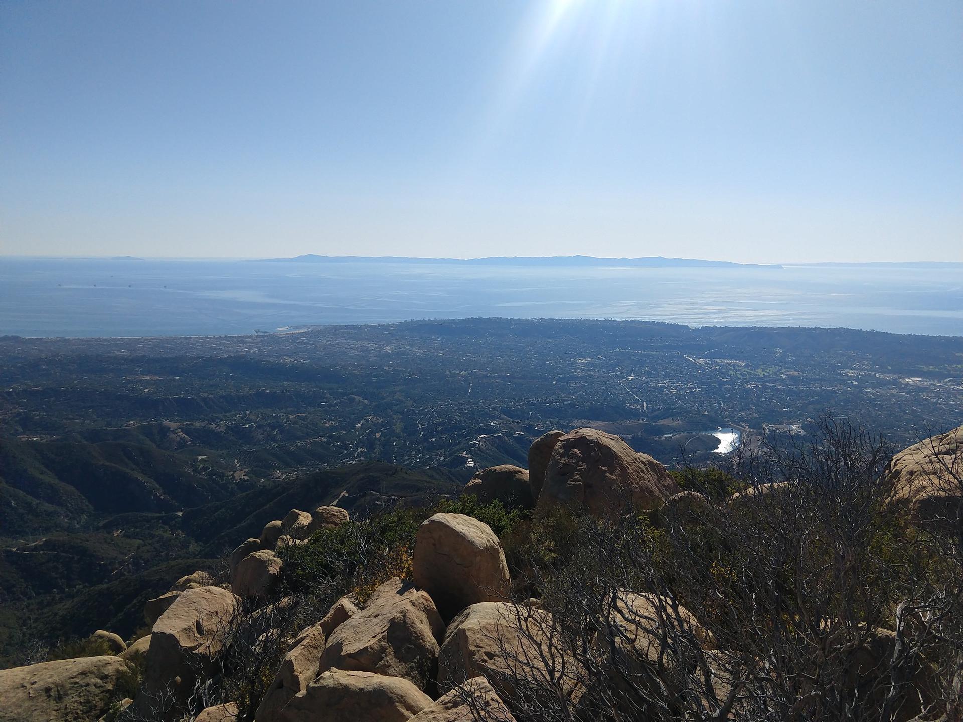 Sunny panoramic view from rocky peak overlooking a city, valley, and ocean.
