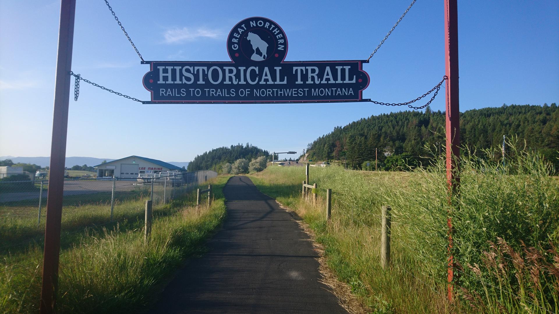 A large sign hangs over a paved trail through grassy fields on a sunny day.