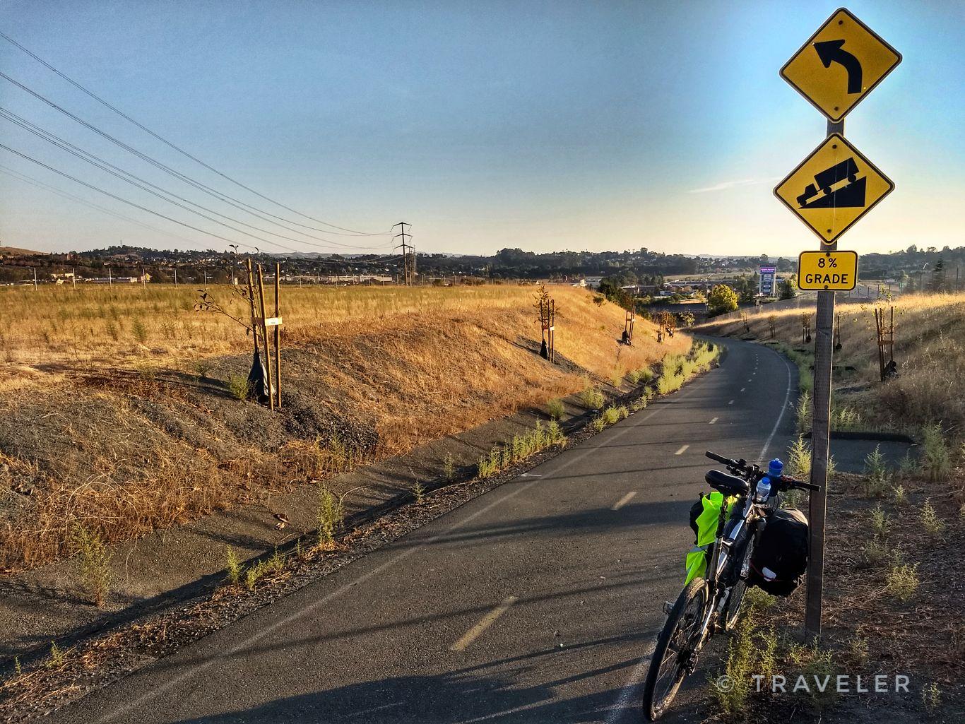 Bicycle parked next to a paved path with warning signs, dry hills.