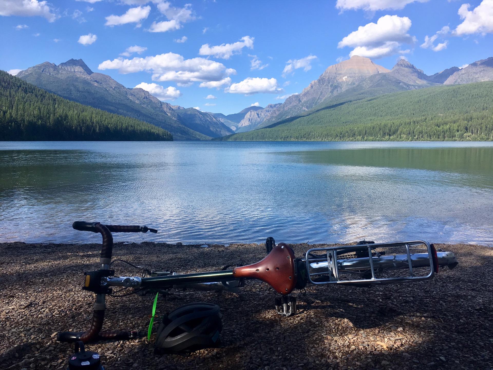 A bicycle and helmet on a rocky shore, with a lake, mountains, and bright sky.