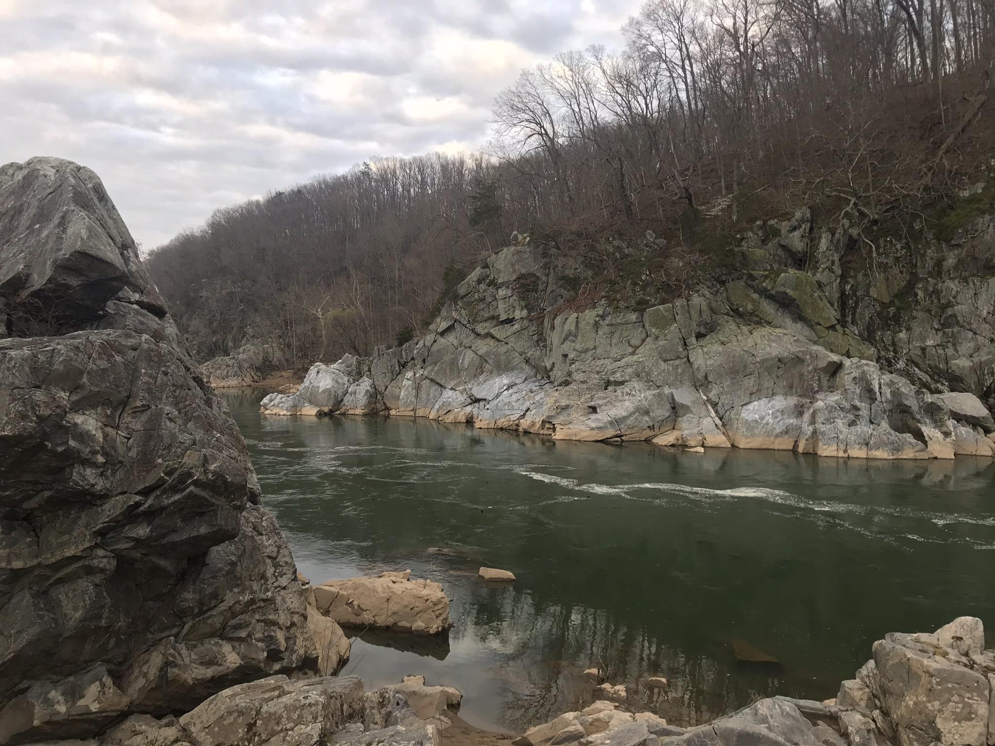 Dark river flowing past large rocks and bare trees under a cloudy sky.
