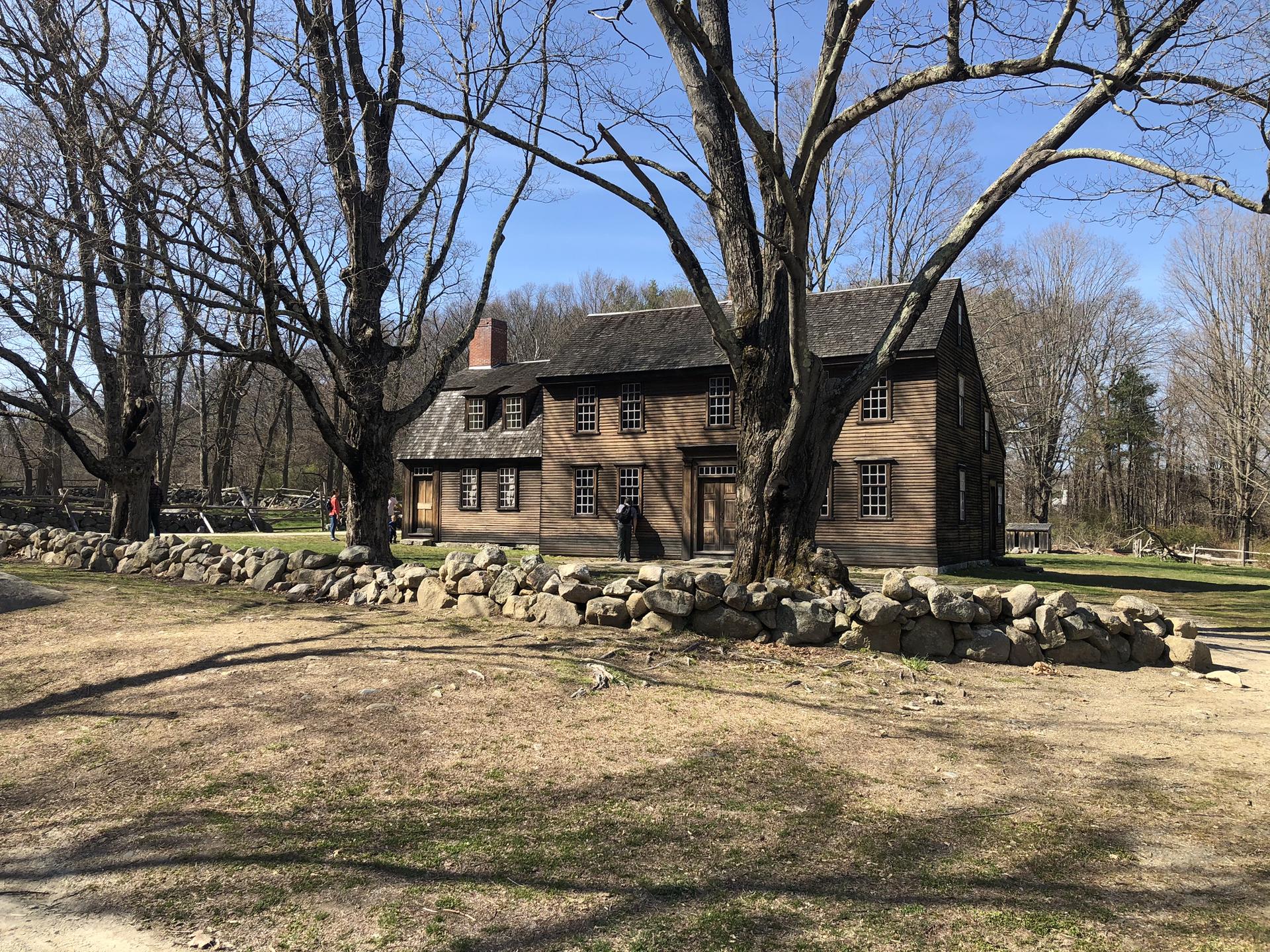 Historic wooden house behind a stone wall, large bare trees, sunny day.