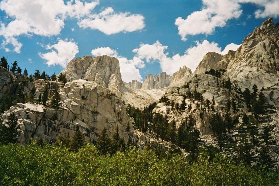 Towering rocky mountains with pine trees and green brush under a cloudy sky. - Whitney Portal Road