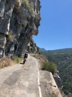 Person cycling on a narrow mountain road beside a cliff and valley under clear sky.