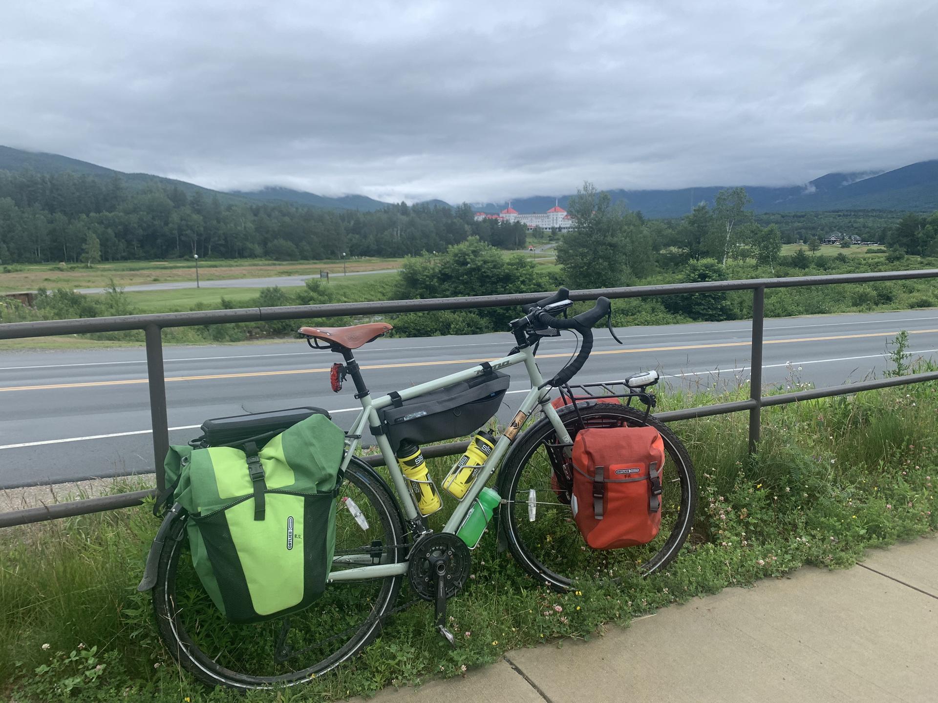 Bicycle with green and orange bags parked by a road, mountains in distance.