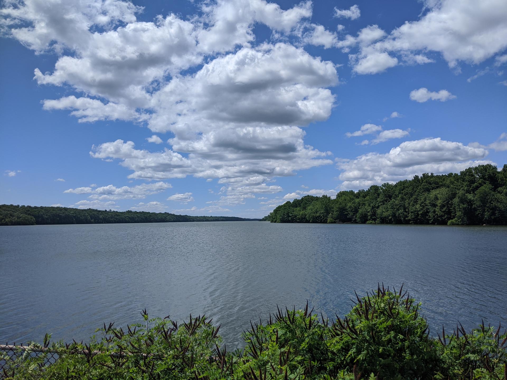 Wide lake surrounded by green forests under a blue, cloudy sky.