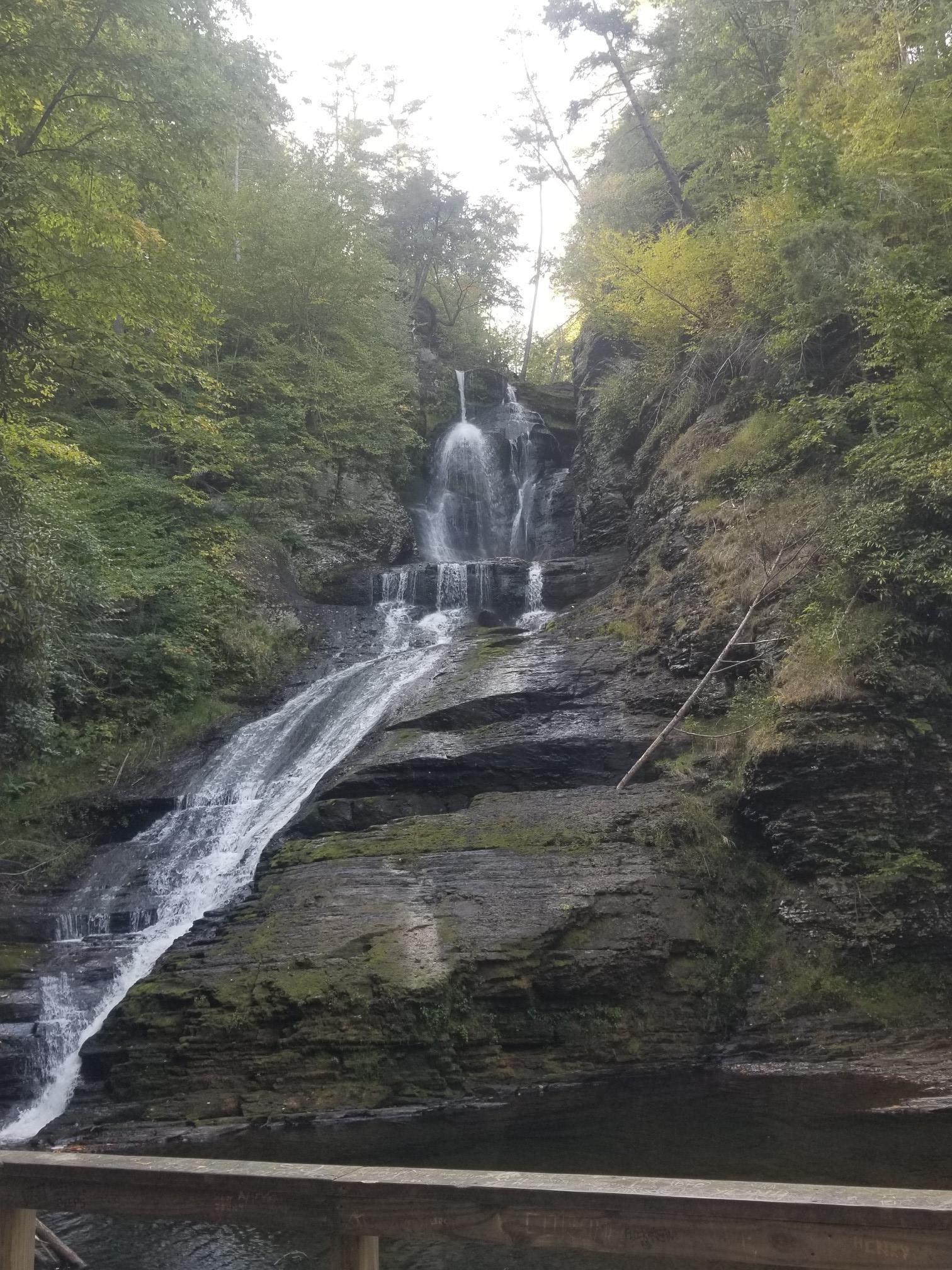 Tall waterfall flows down a rocky, forested slope into a pool, bright light above.