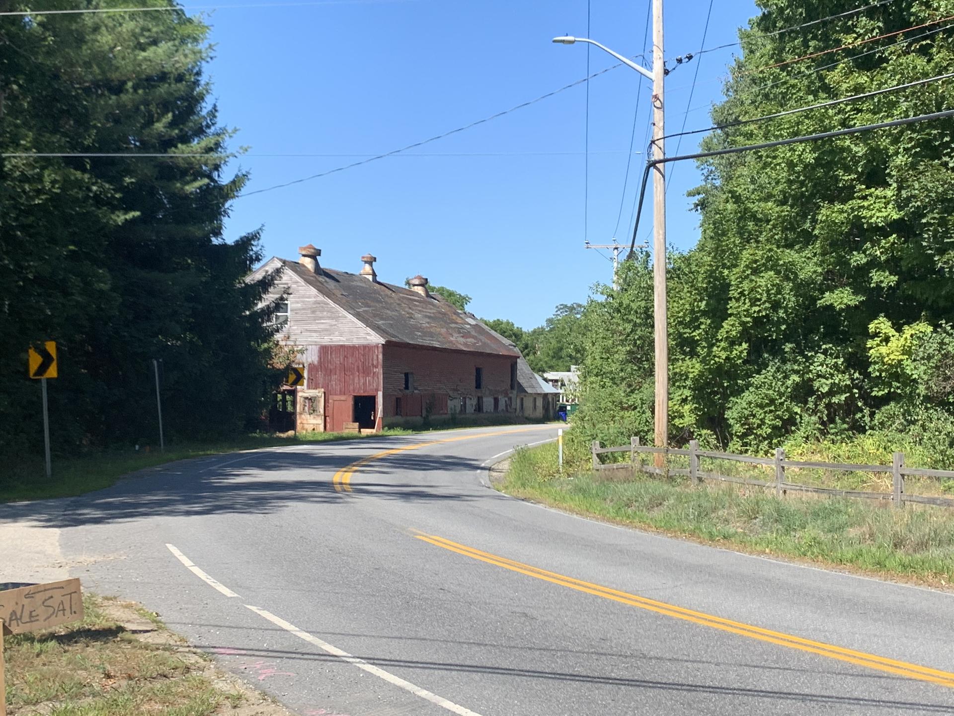 Old red barn beside a paved road with trees under a bright blue sky.