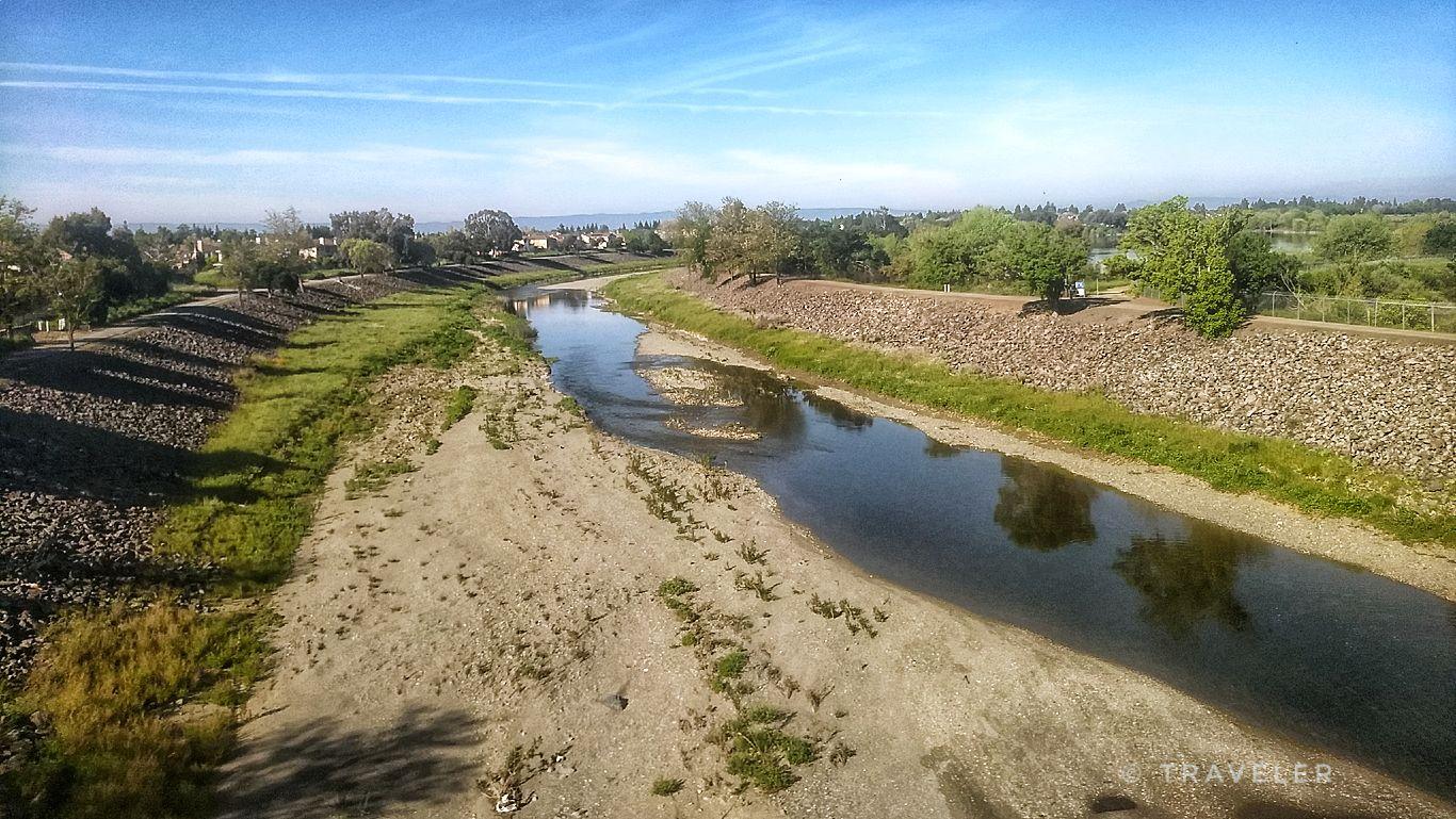 A river with wide, dry banks flowing through a green landscape under a bright sky.