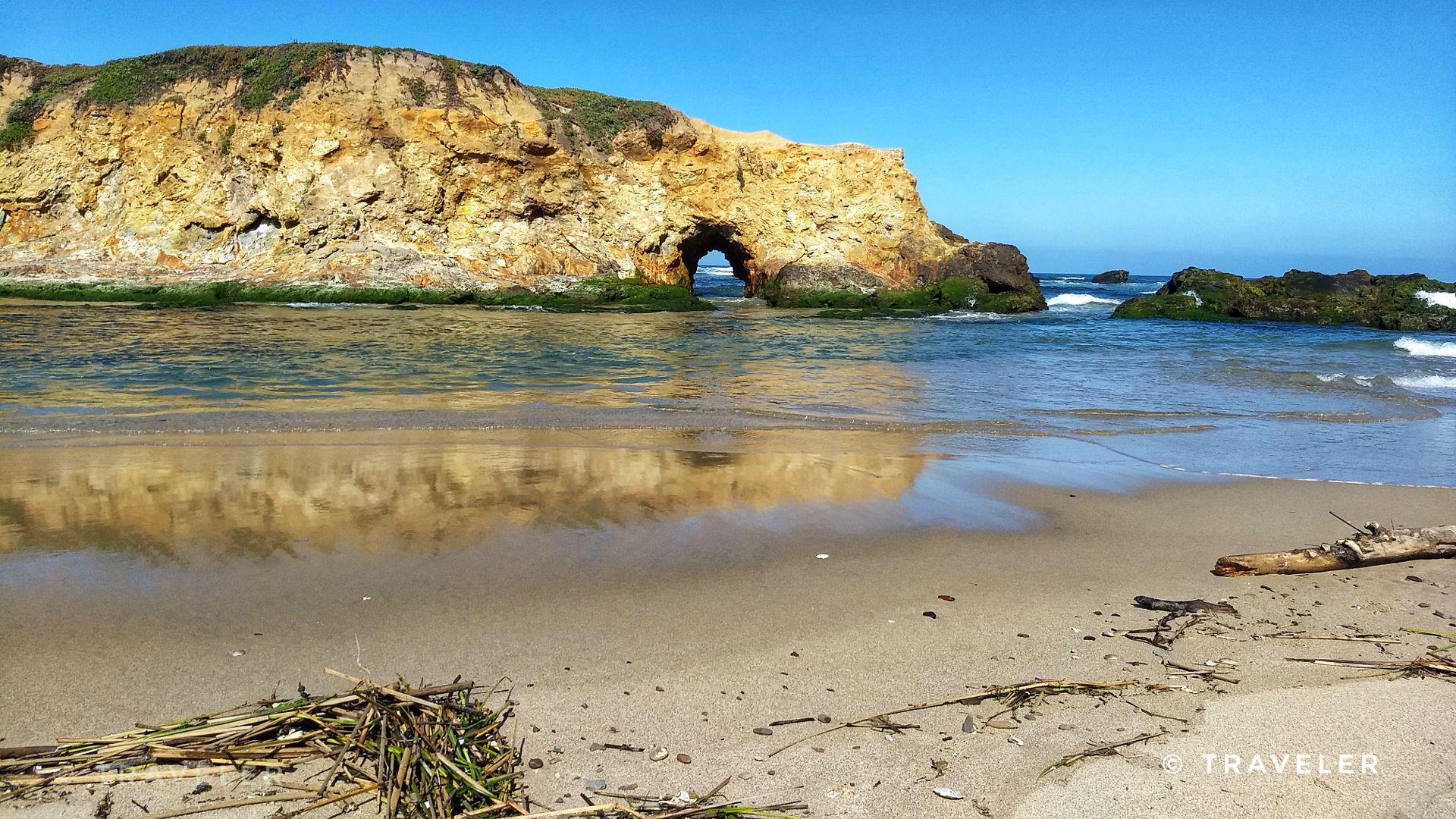 Sandy beach with a rocky sea arch and ocean waves under clear sky.