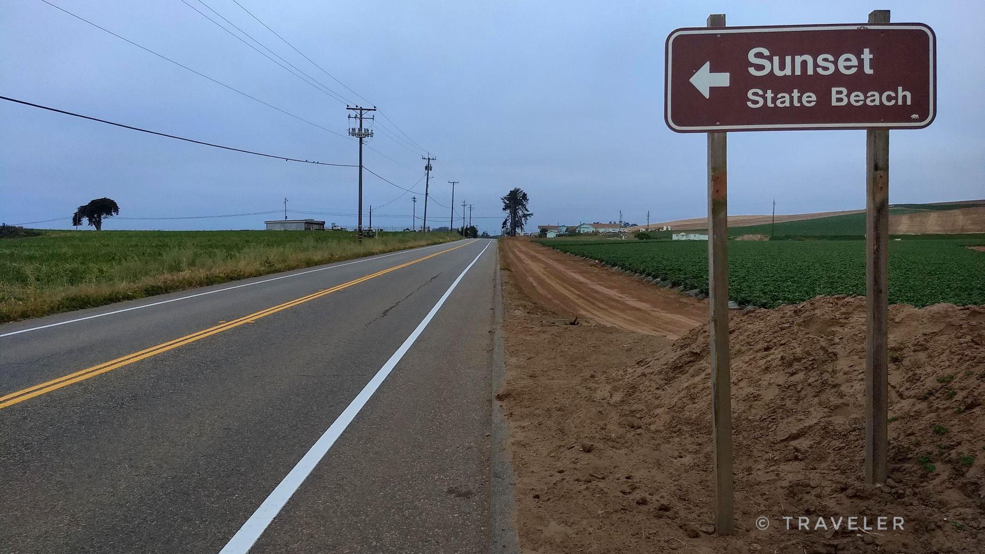 A brown sign with an arrow stands by a paved road and green fields.