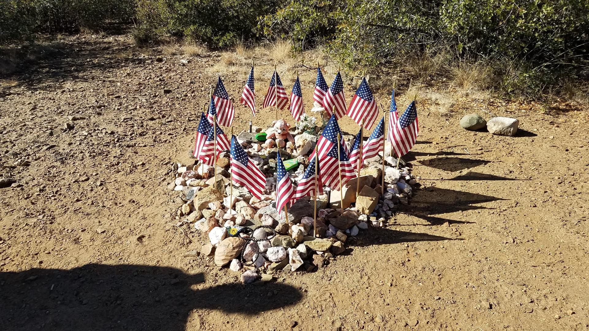 Small American flags and rocks form a heart on dry ground, sunny day.