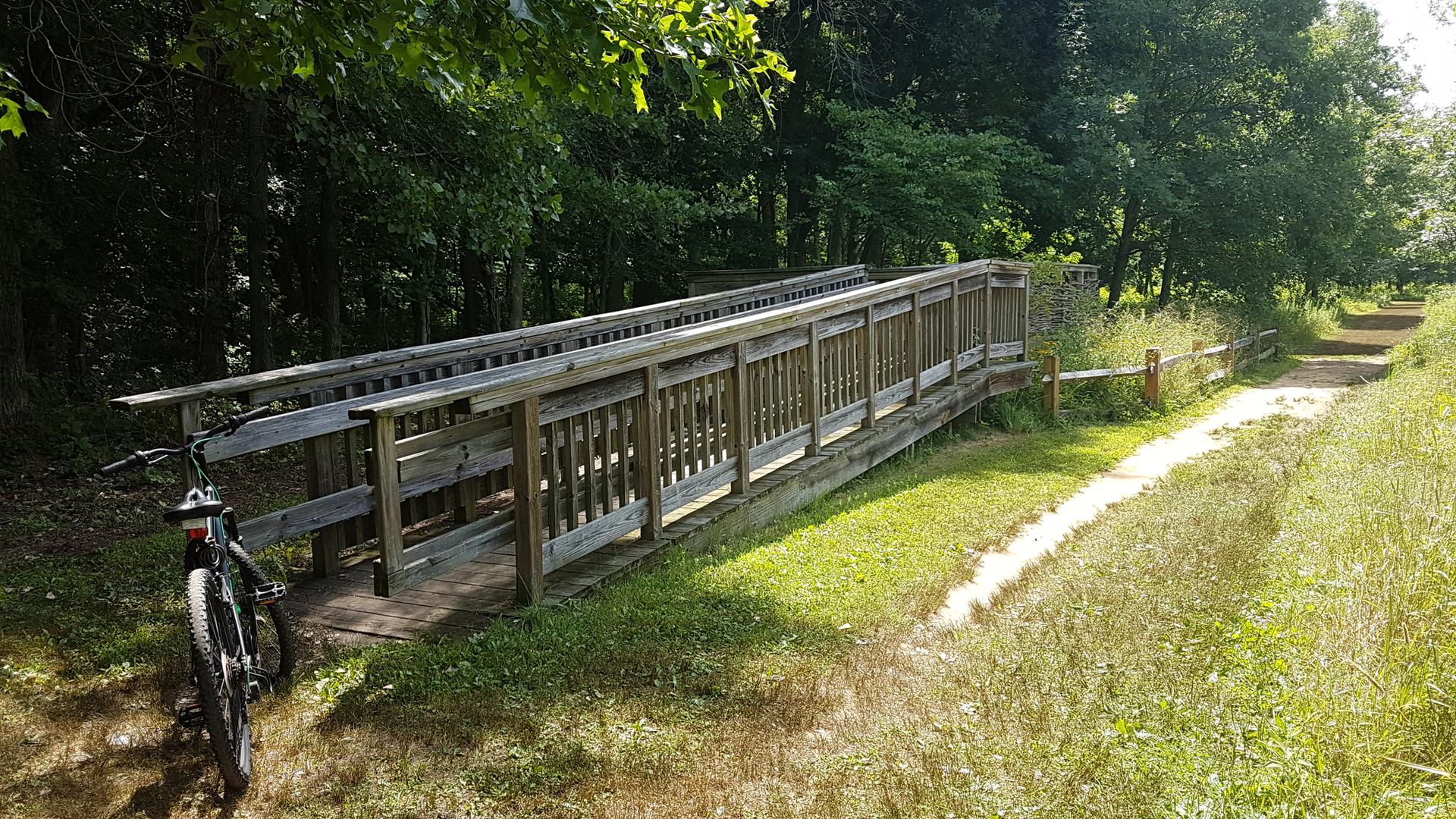 A bicycle rests beside a wooden bridge on a sunny, grassy path.