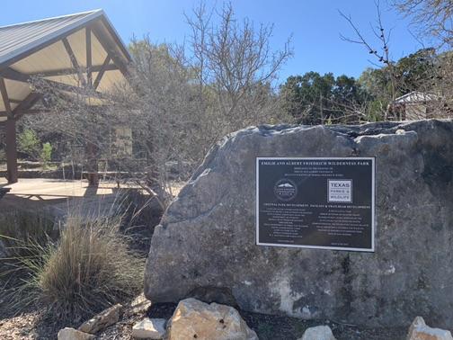 A large rock with a dark plaque in front of a wooden pavilion. Sunny.