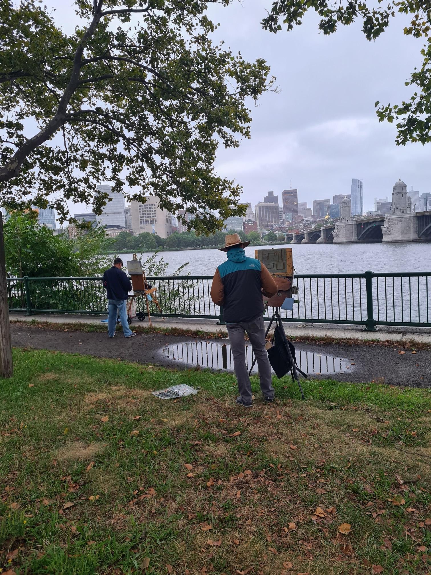 Two artists paint by a river, with a city skyline in the distance.