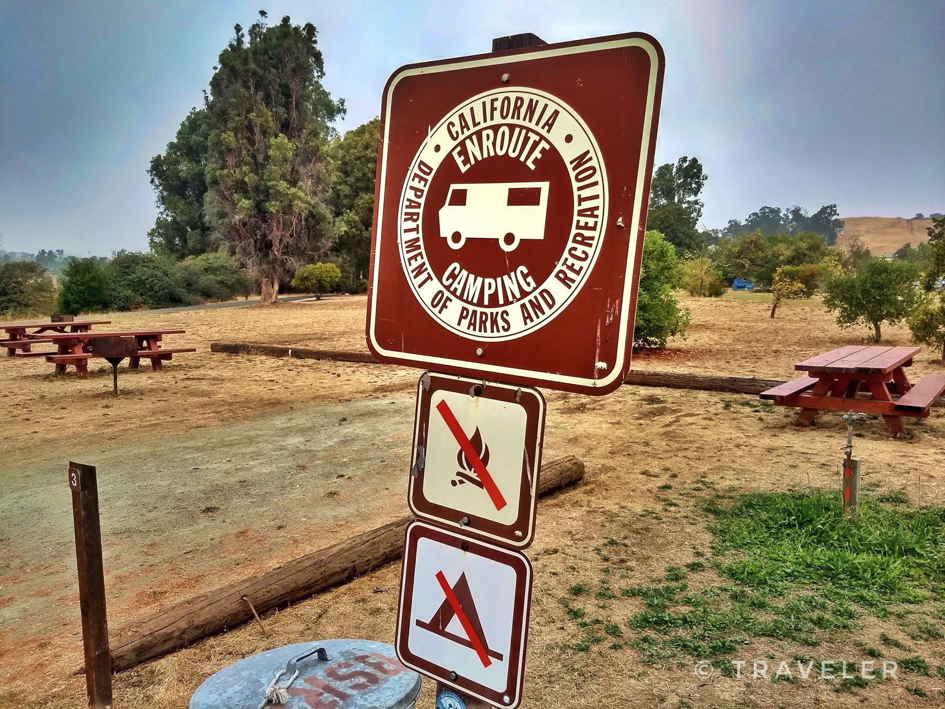 Camping and no-fire signs stand near picnic tables in a dusty, wooded area.