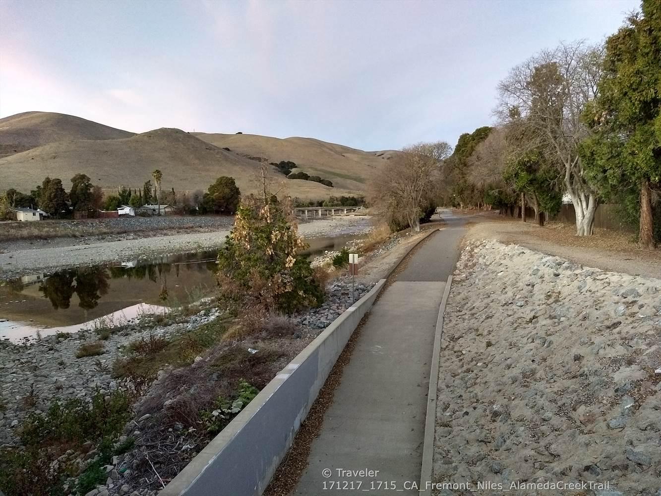 Paved path alongside a riverbed with distant hills under soft light.