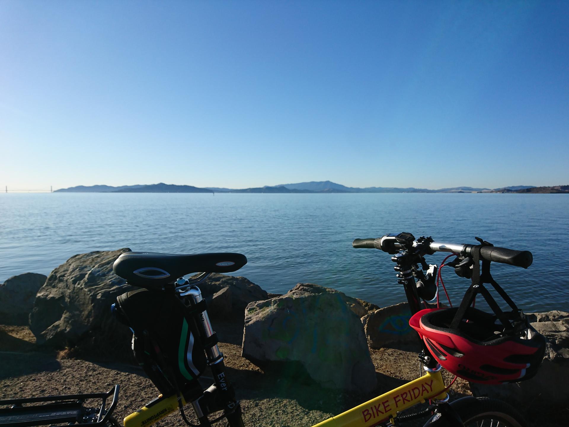 Two bicycles parked on a rocky shore overlooking a calm, sunny water body.