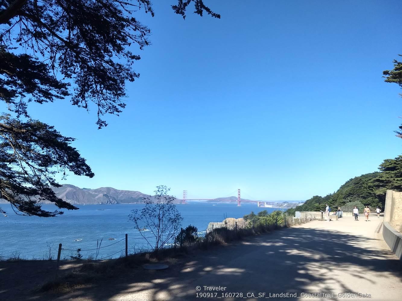 Distant red bridge over water, path with people, trees, clear sky.
