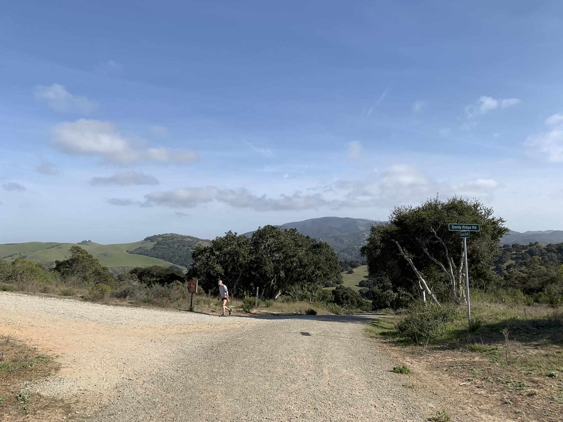 Person walking on a dirt road through hilly, green landscape under blue sky.