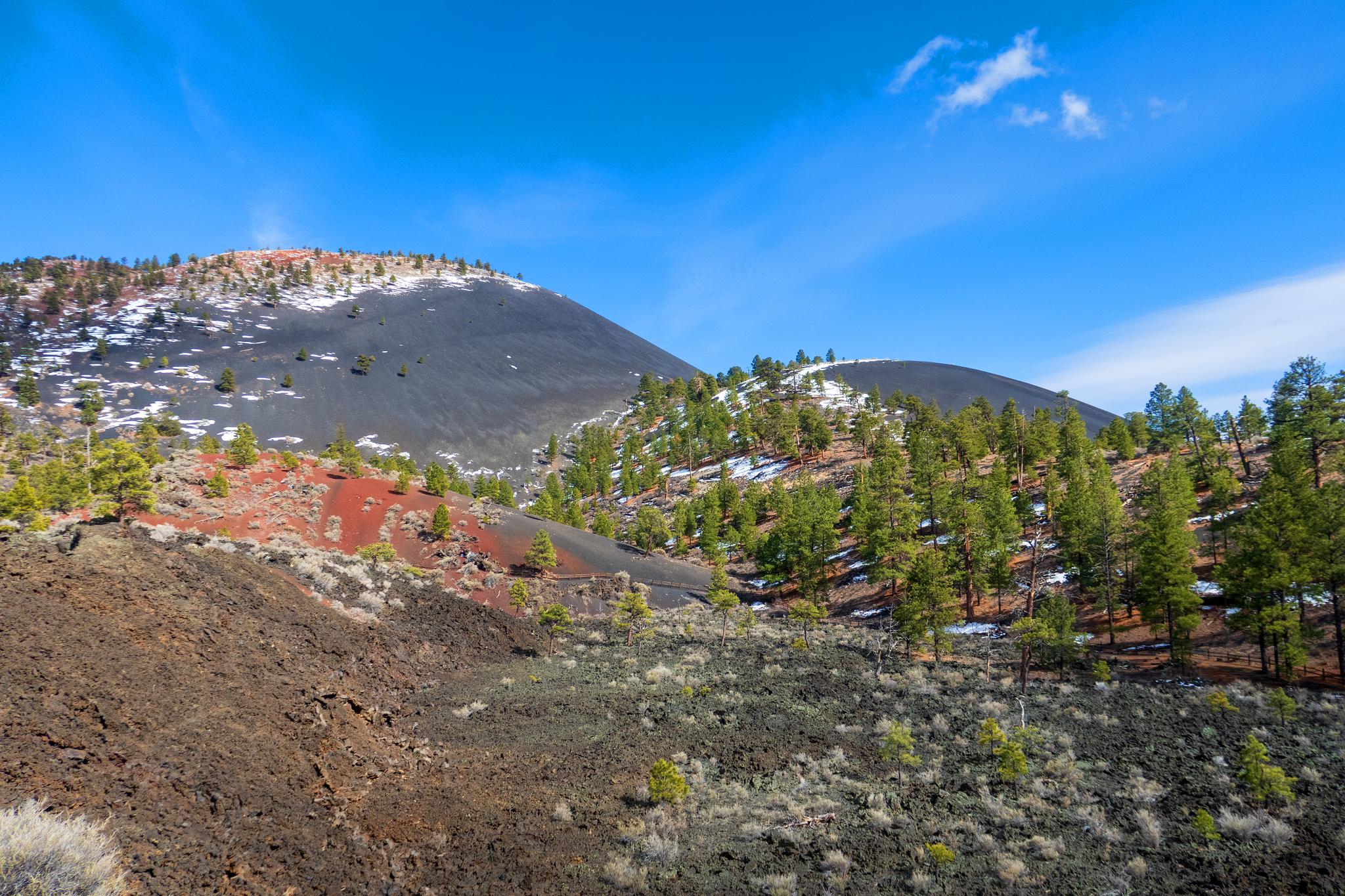 Snow-dusted volcanic hills, red soil, and green pines under a bright blue sky.