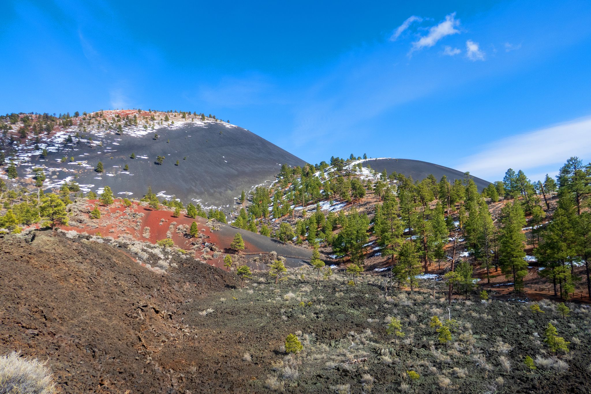 Snow-dusted volcanic hills, red soil, and green pines under a bright blue sky. - Lenox Crater and Lava Flow Trail Loop