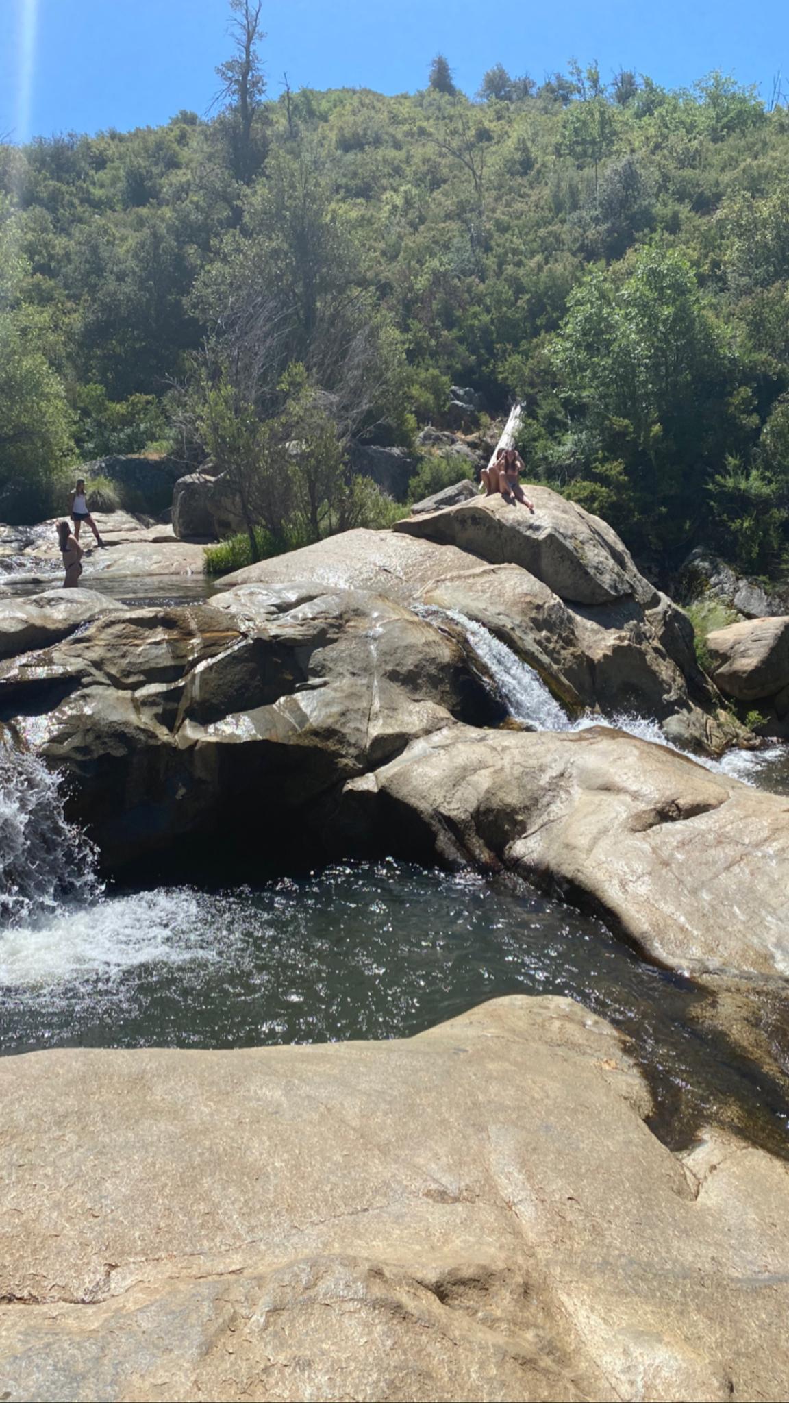 People relax on sunlit rocks beside a flowing river and small waterfall.